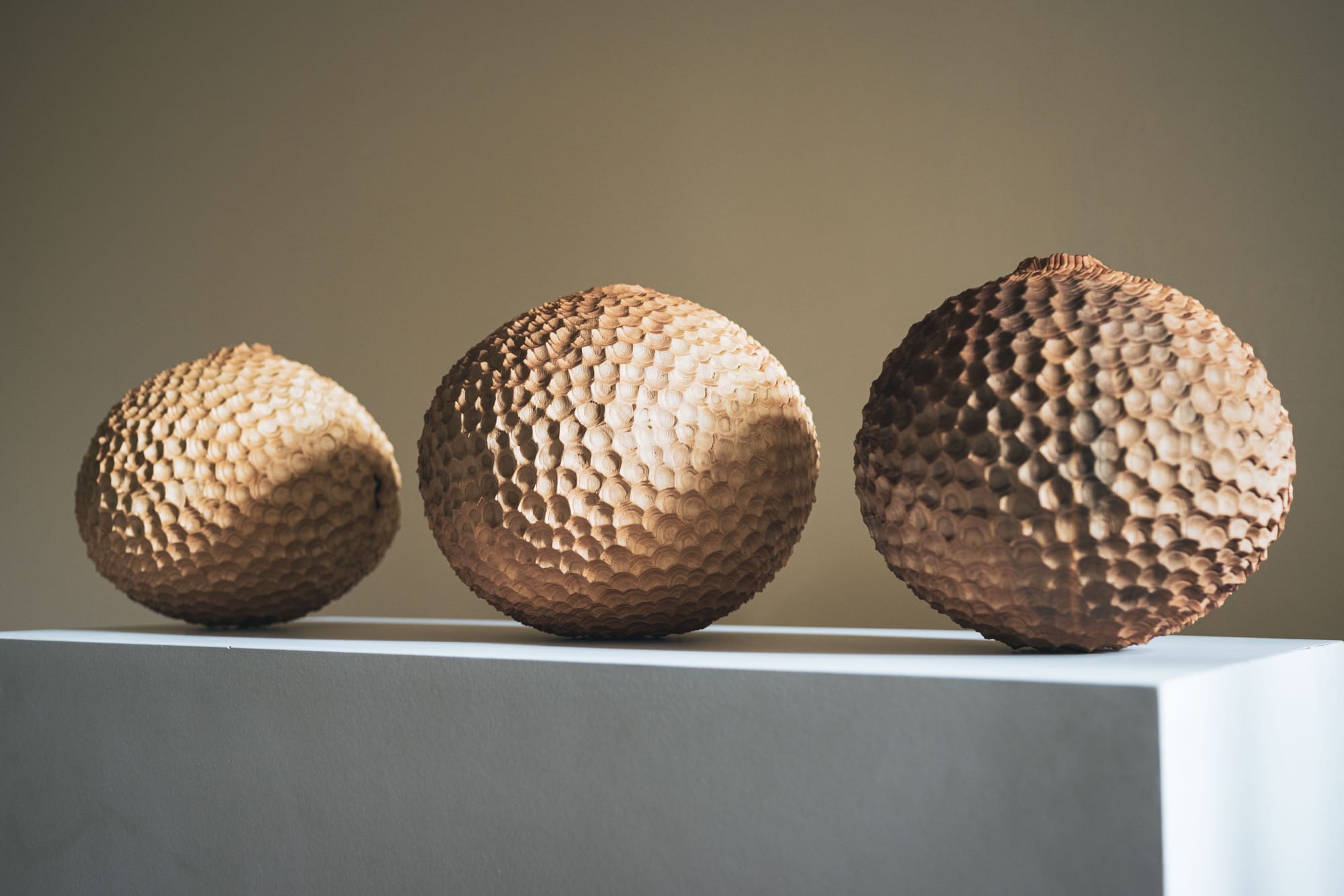 a series of three handmade wooden vessels with faceted surfaces, situated on a pedestal against a neutral background