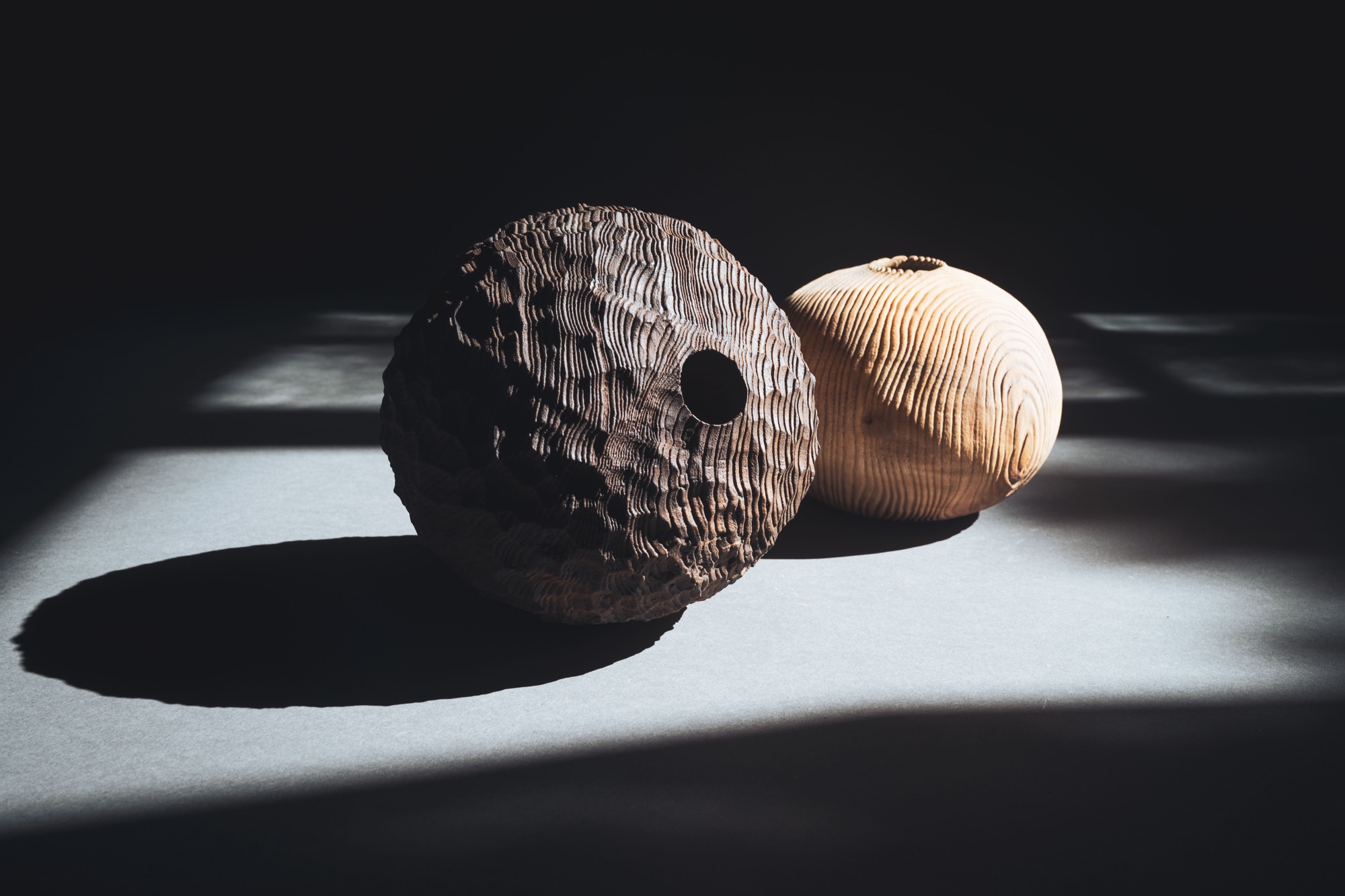 two handmade wooden vessels with textured surfaces, illuminated against a dark background