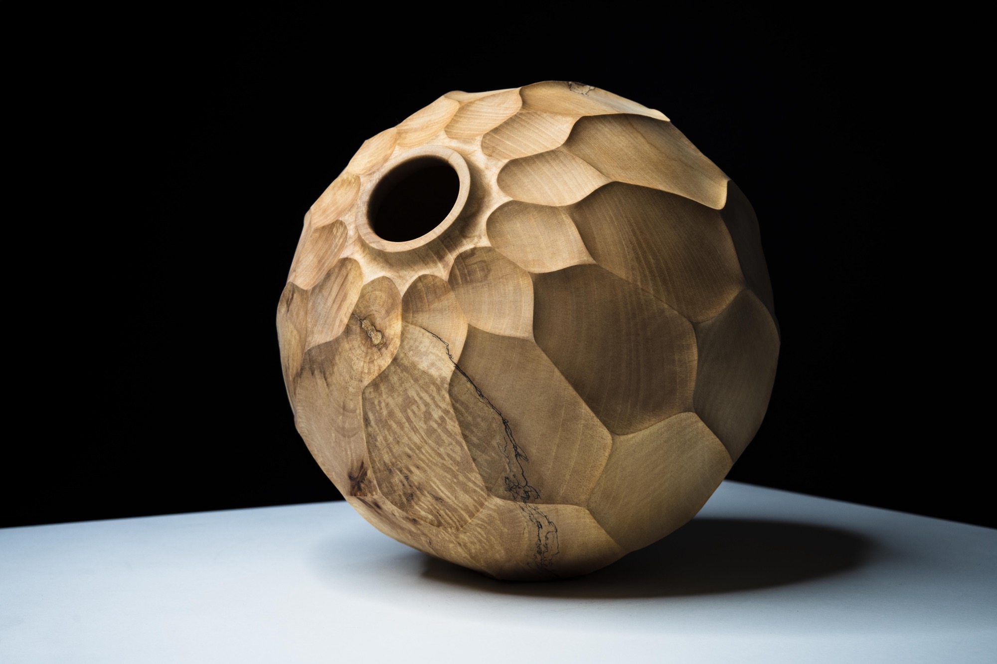 a handmade wooden vessel with a faceted surface, illuminated against a dark background