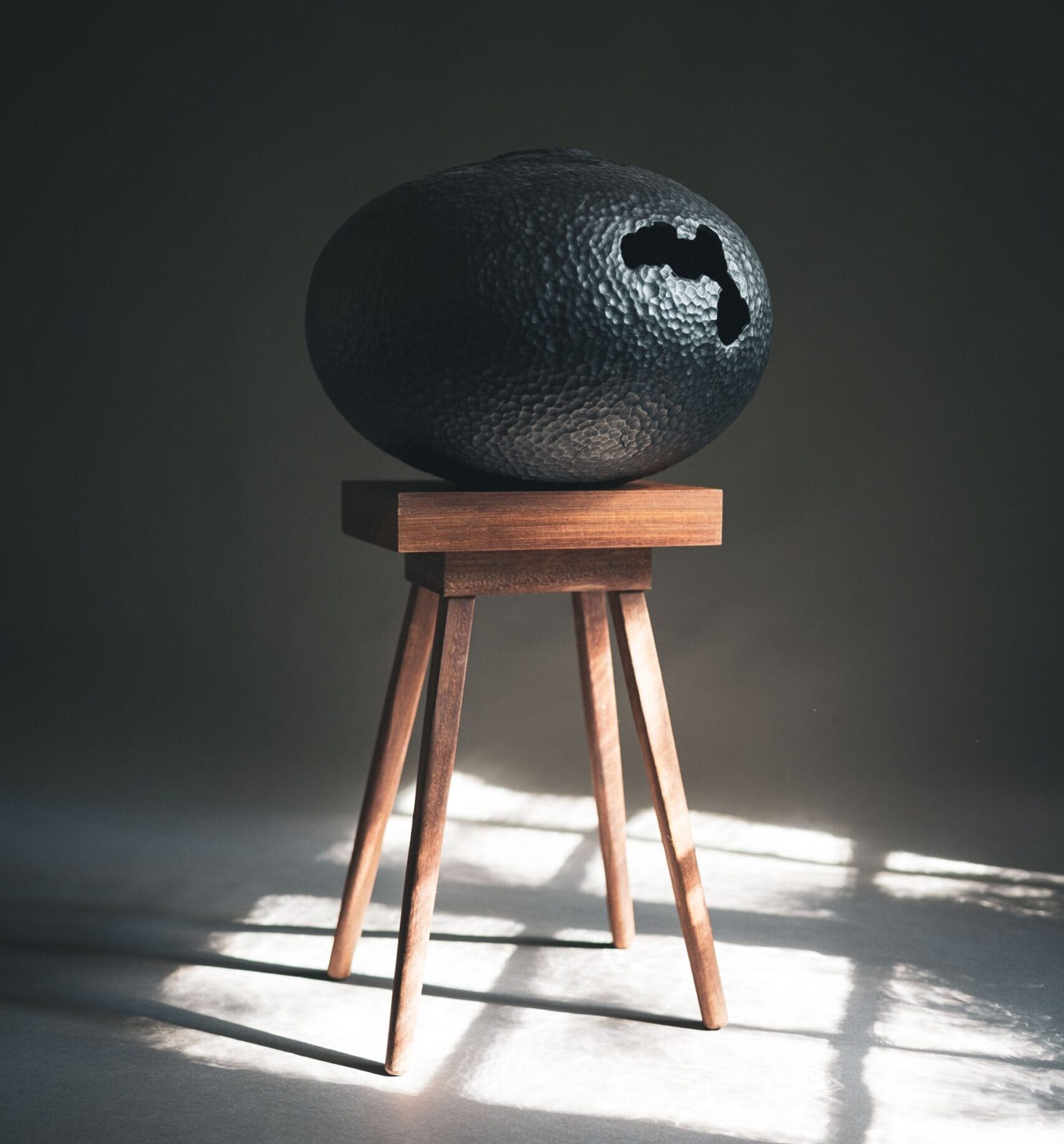 a handmade black wooden vessel sitting on a wooden stool, illuminated against a dark background