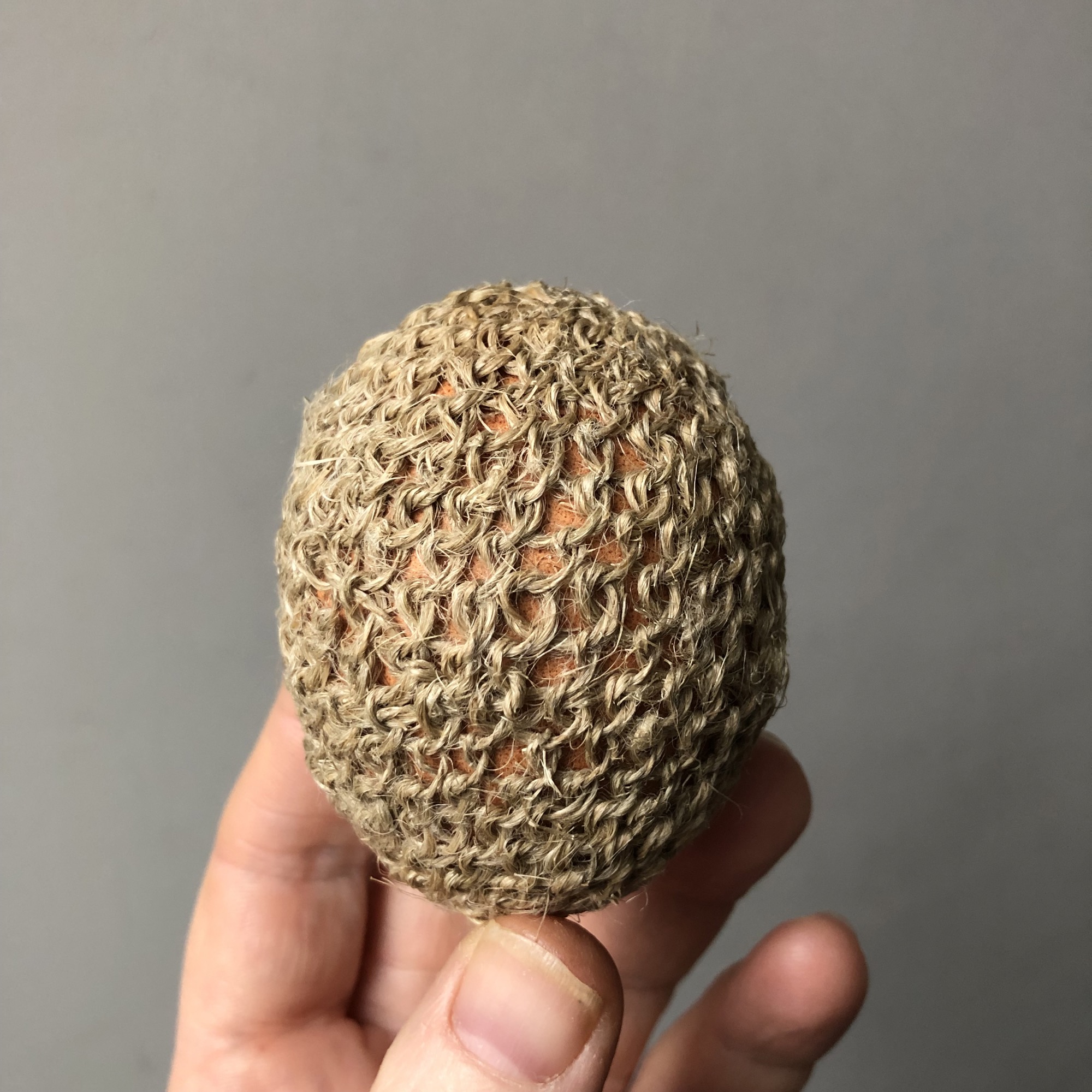 a hand holds a brown hen egg that has been completed enclosed in a woven layer of natural fiber