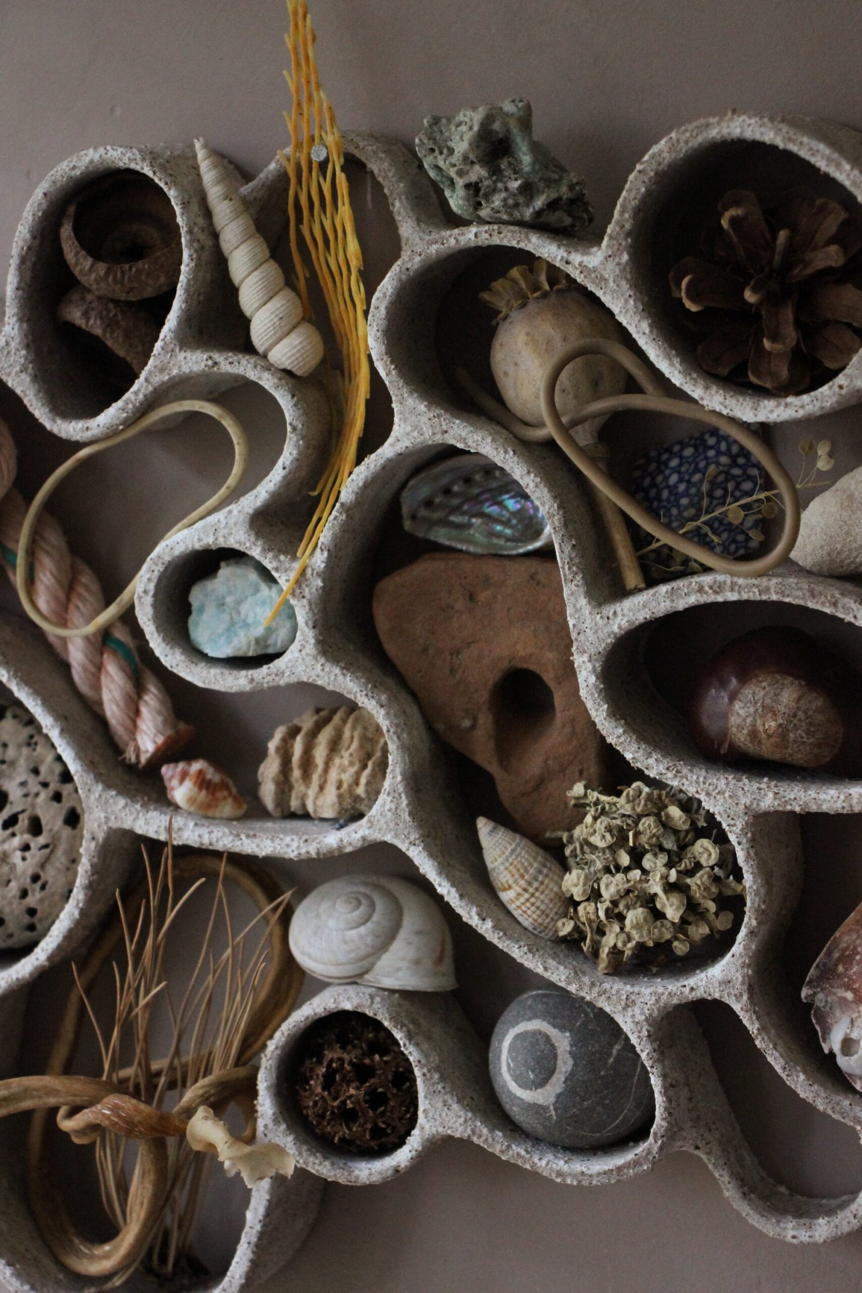 A detail photo of a functional, abstract ceramic sculpture hanging on a dark wall, filled with shells, pebbles, and other found organic items.