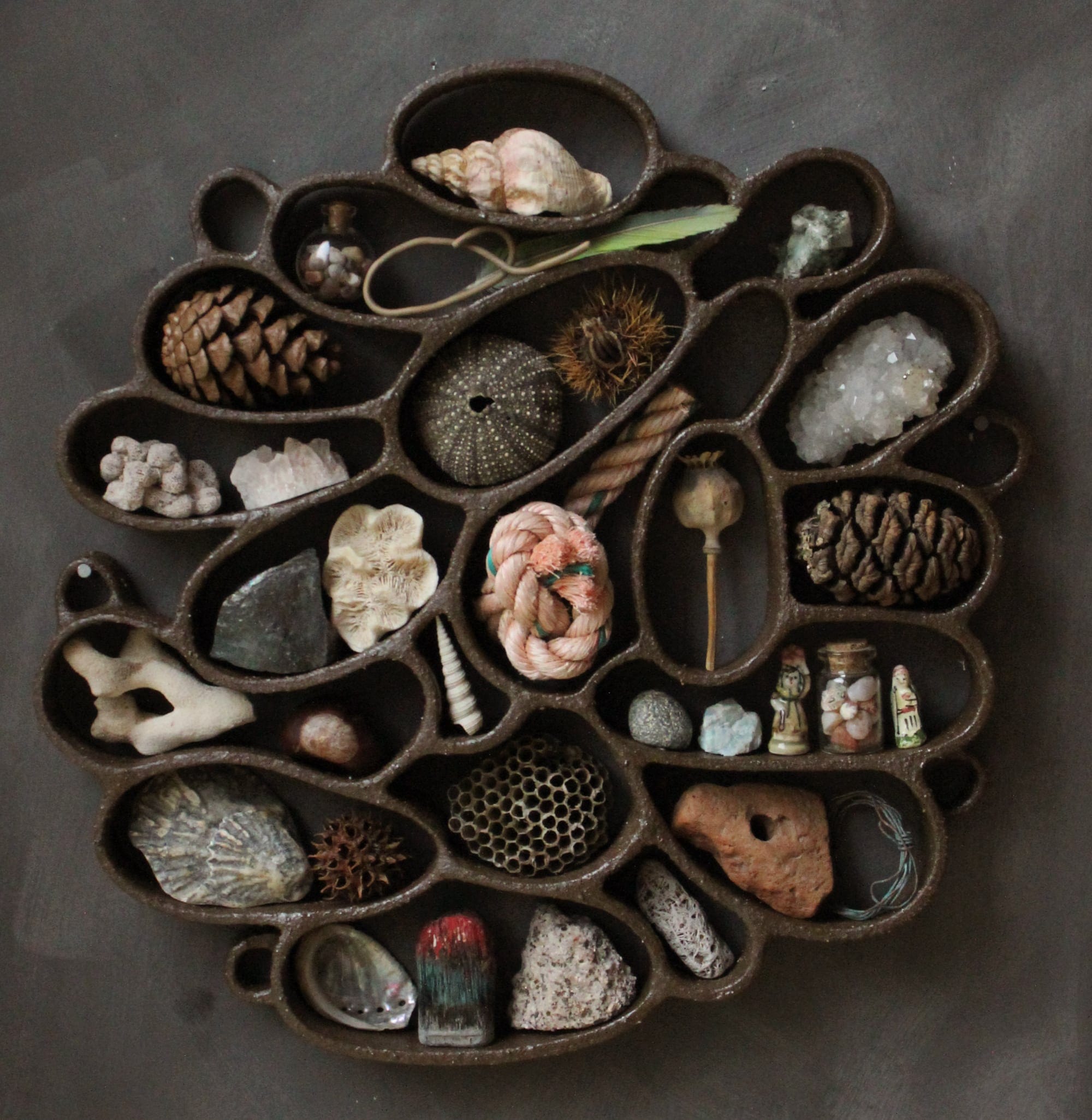 A functional, abstract ceramic sculpture hanging on a dark wall, filled with shells, pebbles, and other found organic items.