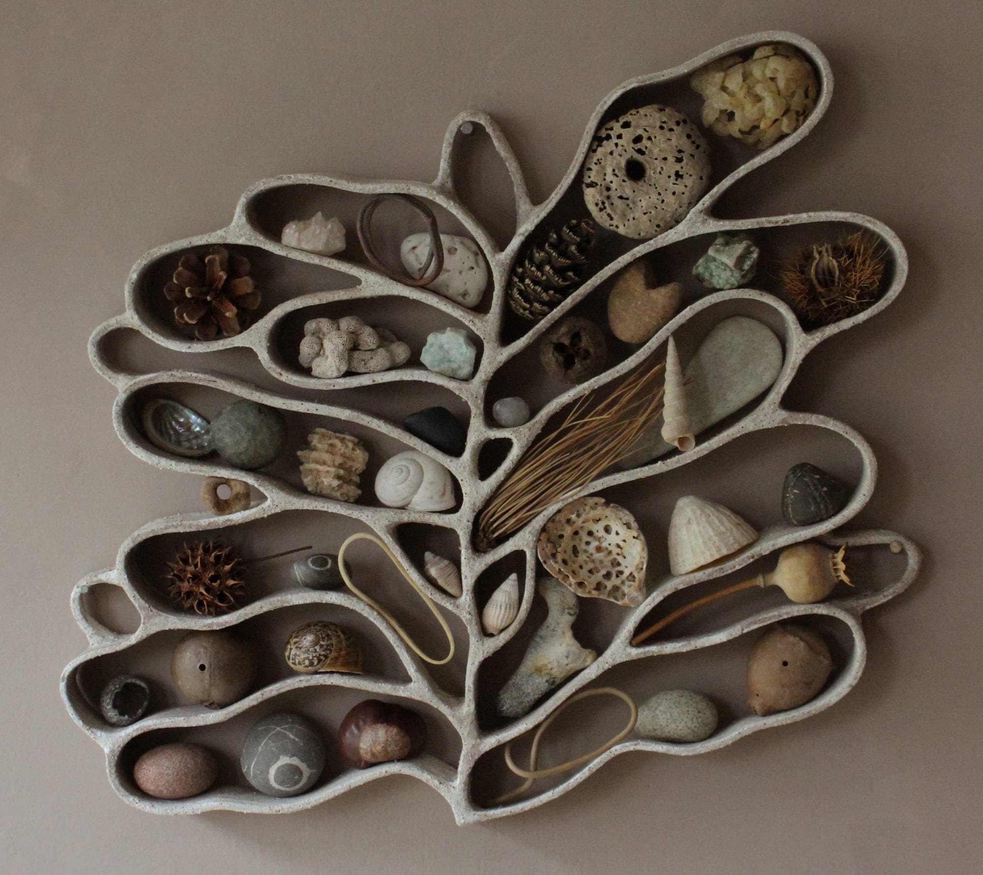 A functional, abstract ceramic sculpture hanging on a beige wall, filled with shells, pebbles, and other found organic items.