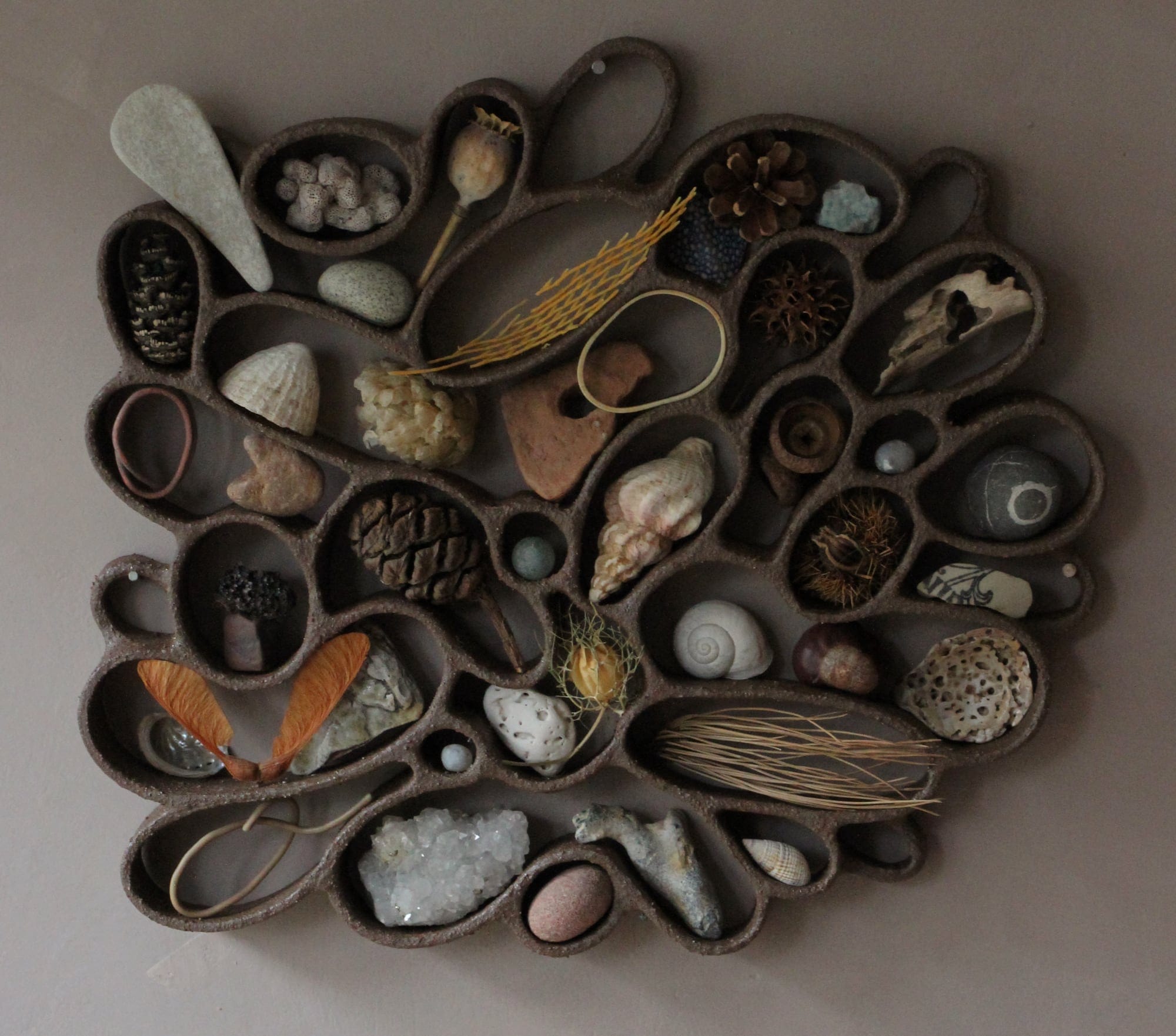 A functional, abstract ceramic sculpture hanging on a dark wall, filled with shells, pebbles, and other found organic items.