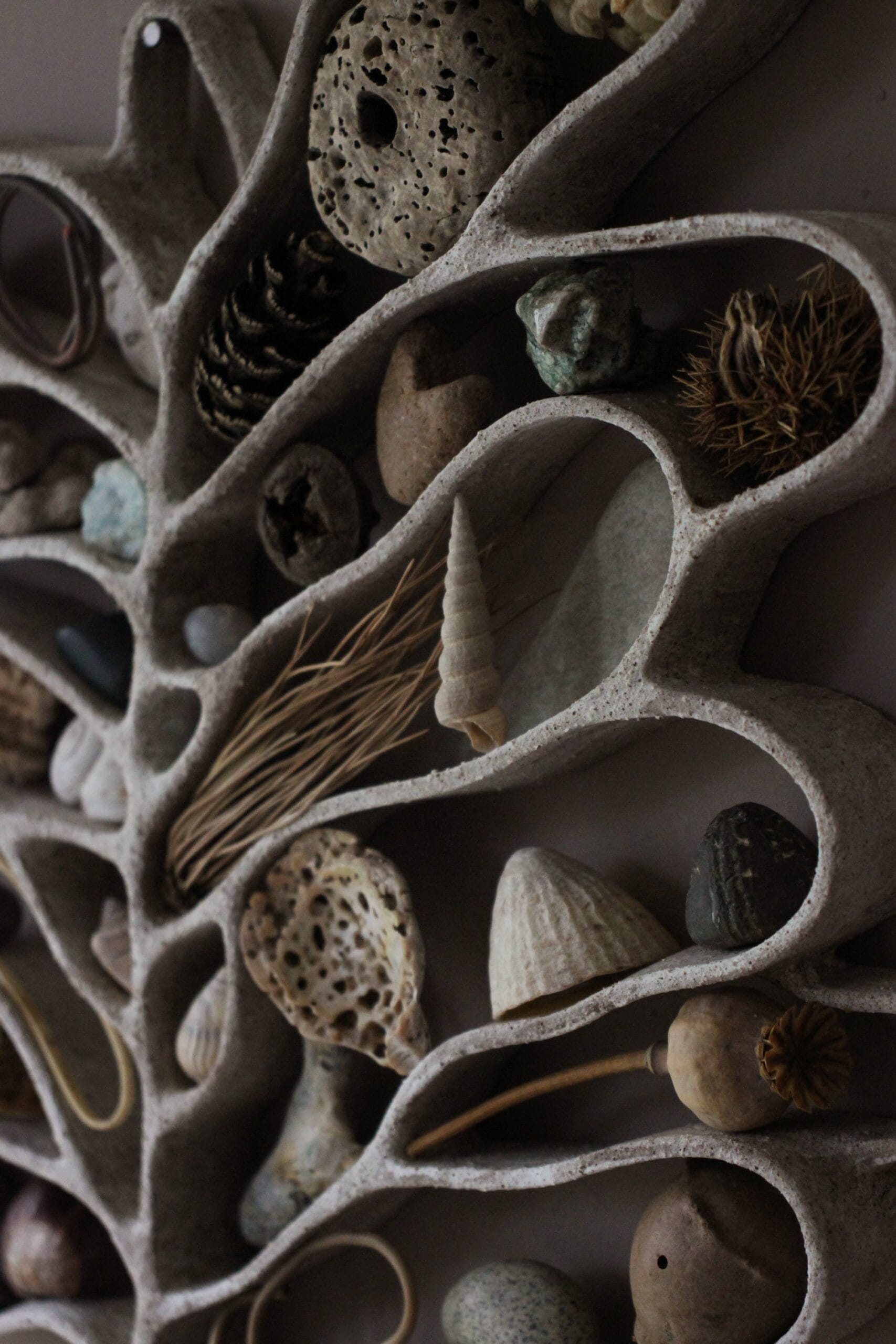 A detail photo of a functional, abstract ceramic sculpture hanging on a dark wall, filled with shells, pebbles, and other found organic items.