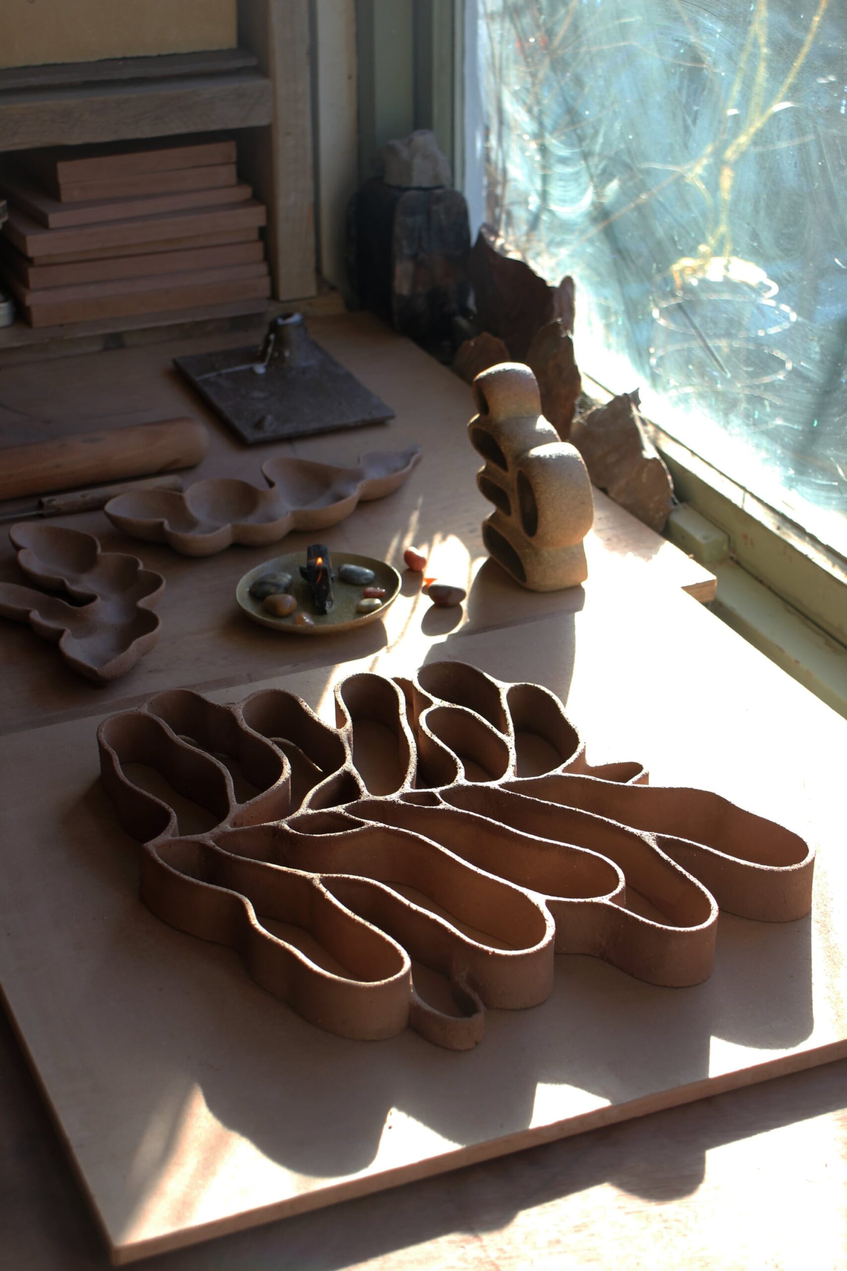 a ceramic sculpture in the shape of a leaf rests on a tabletop by a window with other small ceramic platters and sculptures nearby