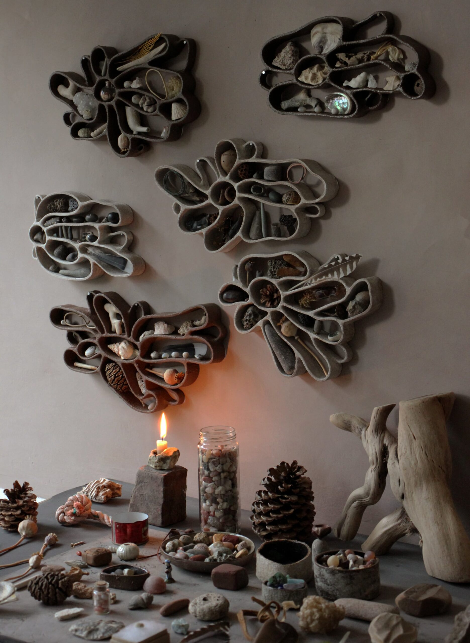 Several functional, abstract ceramic sculptures hang on a dark wall, filled with shells, pebbles, and other found organic items. a tabletop with pinecones, stones, a burning candle, logs, and other natural objects sits below