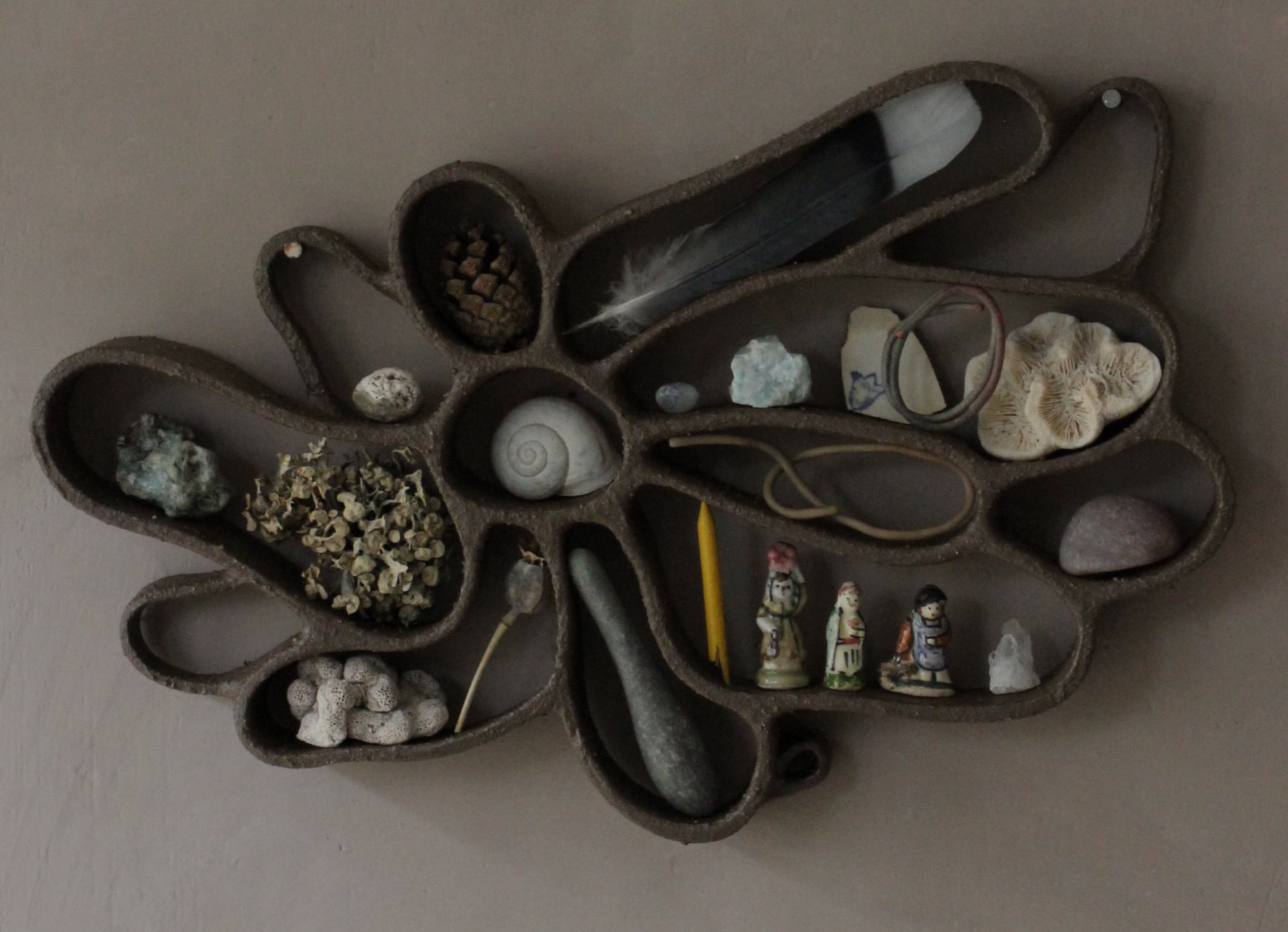 A functional, abstract ceramic sculpture hanging on a dark wall, filled with shells, pebbles, and other found organic items.