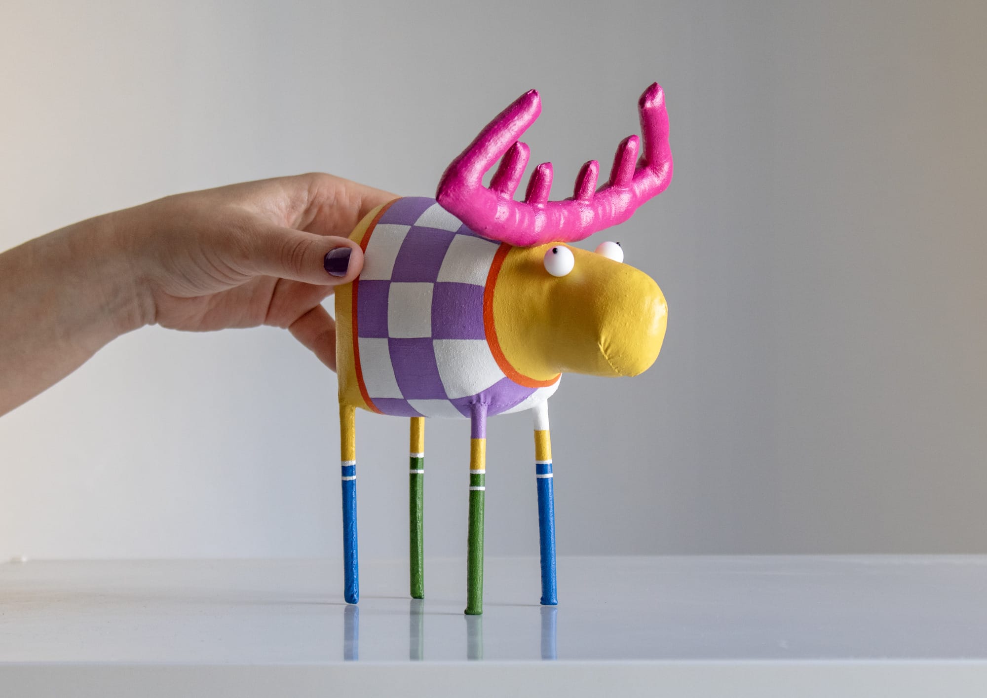 a hand holds up a yellow moose with long, skinny legs. it has pink antlers and wears a purple and white checkered sweater. 