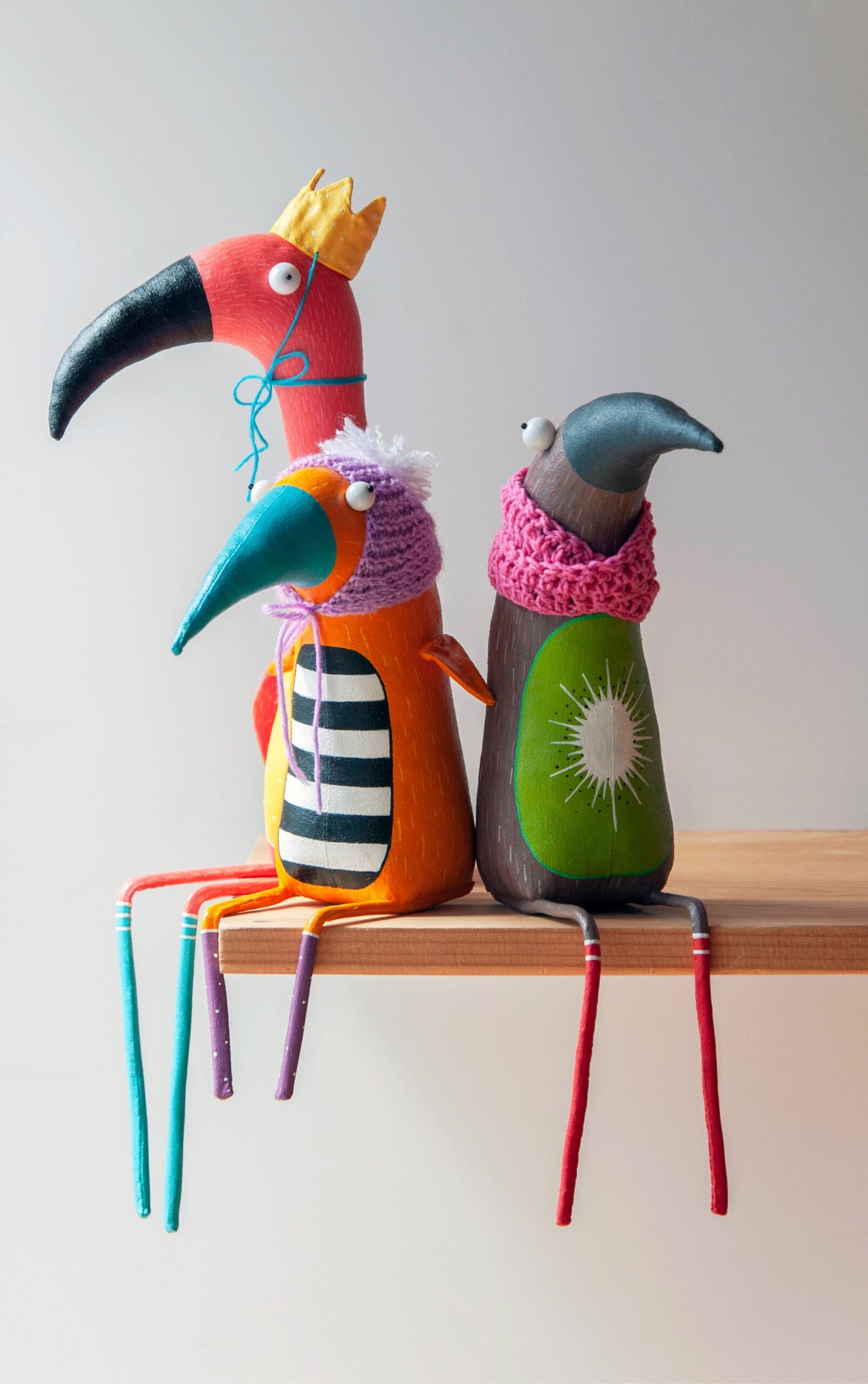 an orange bird with a striped belly and knit bonnet, a tall pink flamingo with a tie-on crown, and a kiwi bird with the fruit on its belly wearing a knit scarf all sit together on a wooden shelf.