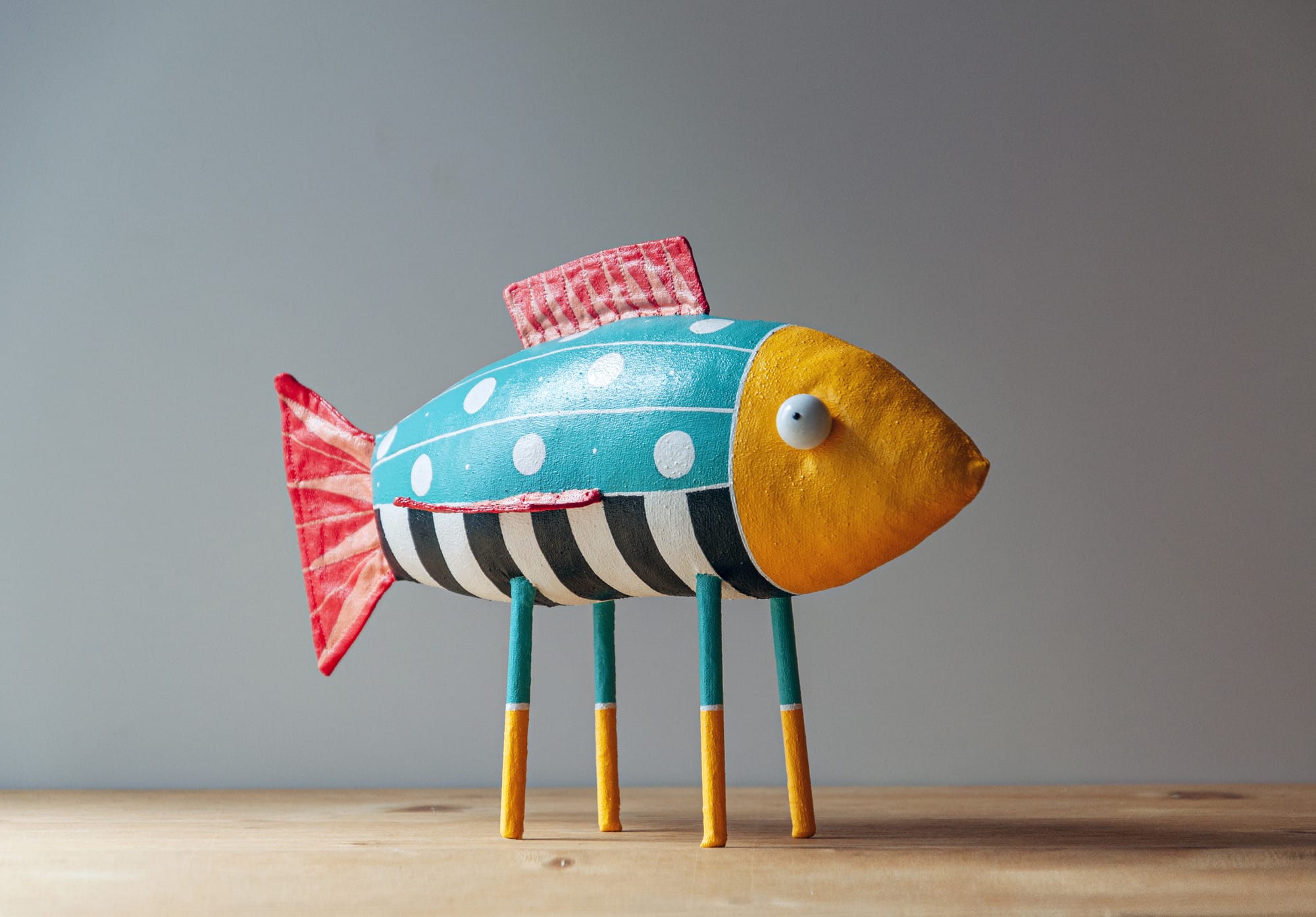 a fish donning primary colors, black and white stripes, and four long, stocky legs stands atop a wooden surface.