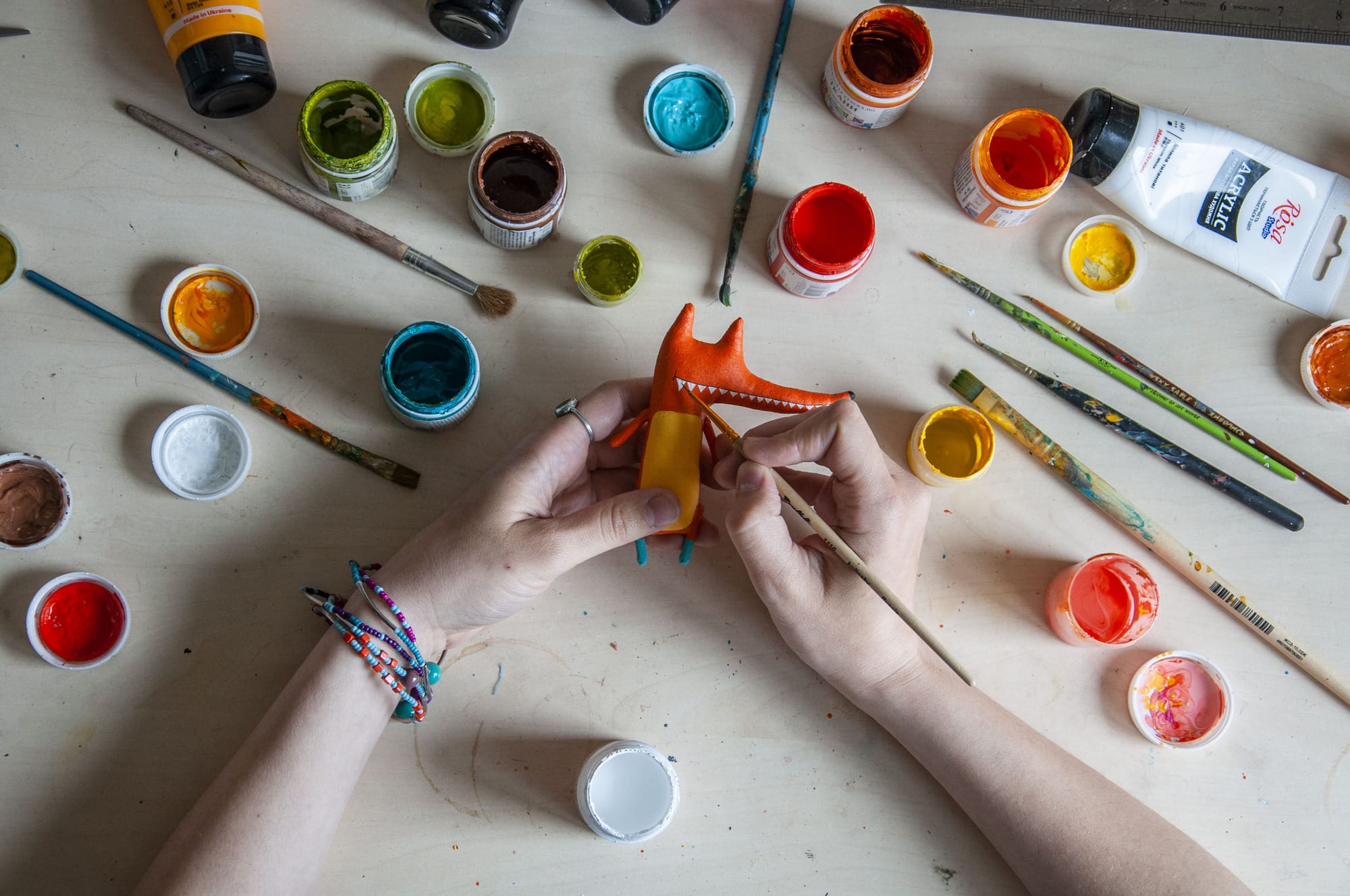 an overhead shot of the artist painting a red fox figure with paint. many jars of paint surround the area.