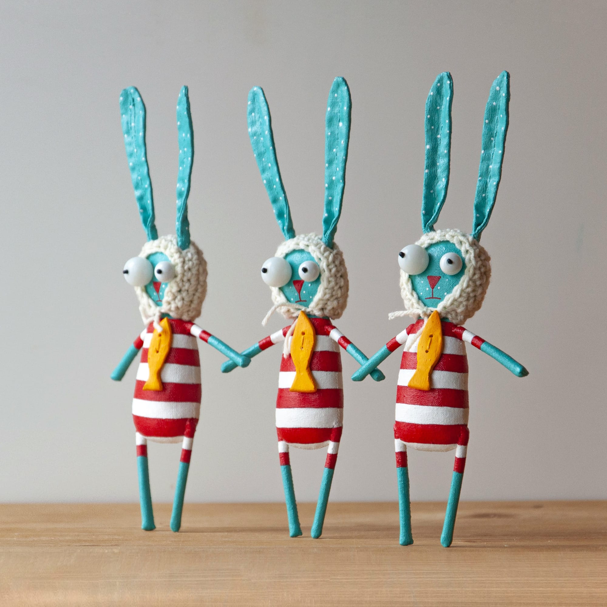 three identical bright blue rabbits wear striped red and white tops, white knit bonnets, and fish brooches.