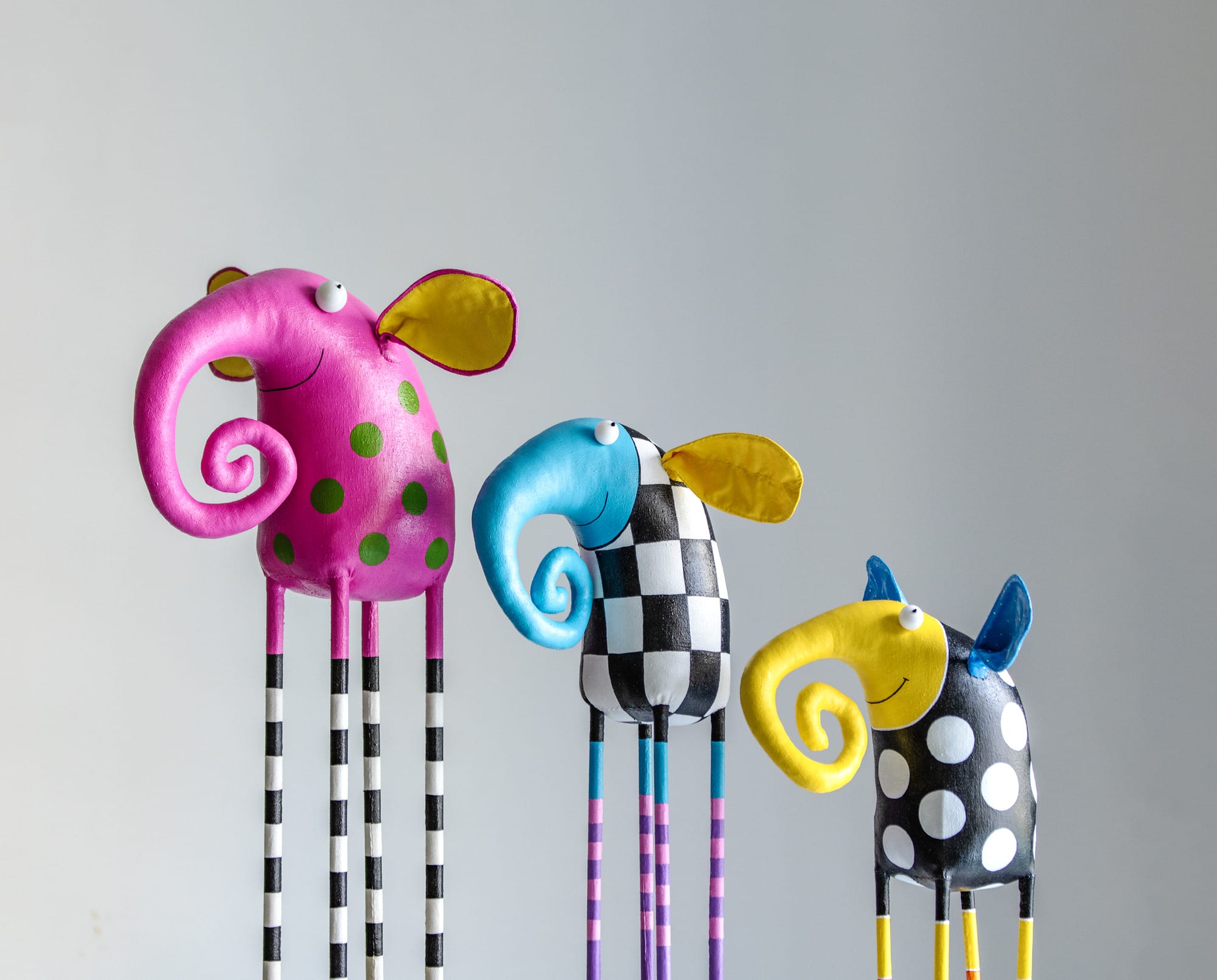 three vibrantly colored elephants with swirly trunks stand in descending height order, each adorned with patterns like stripes, checkered squares, and polka-dots.