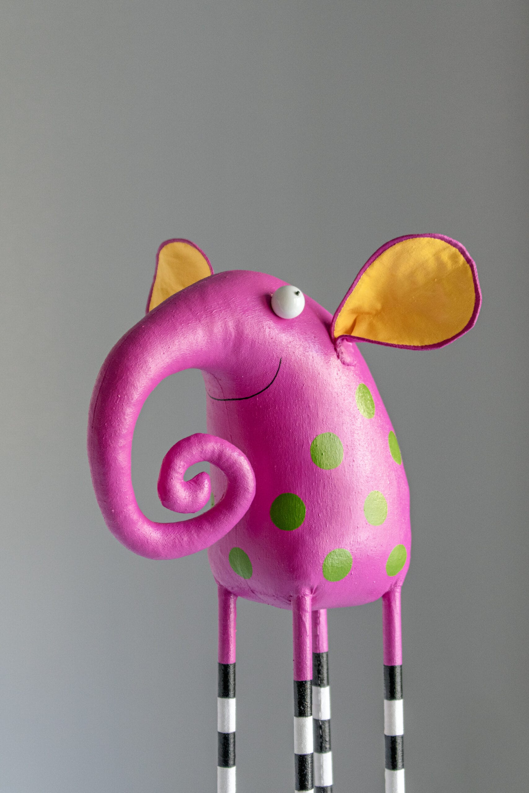 a hot pink elephant with a swirly trunk smiles. it has large yellow ears and long, stocky striped legs.