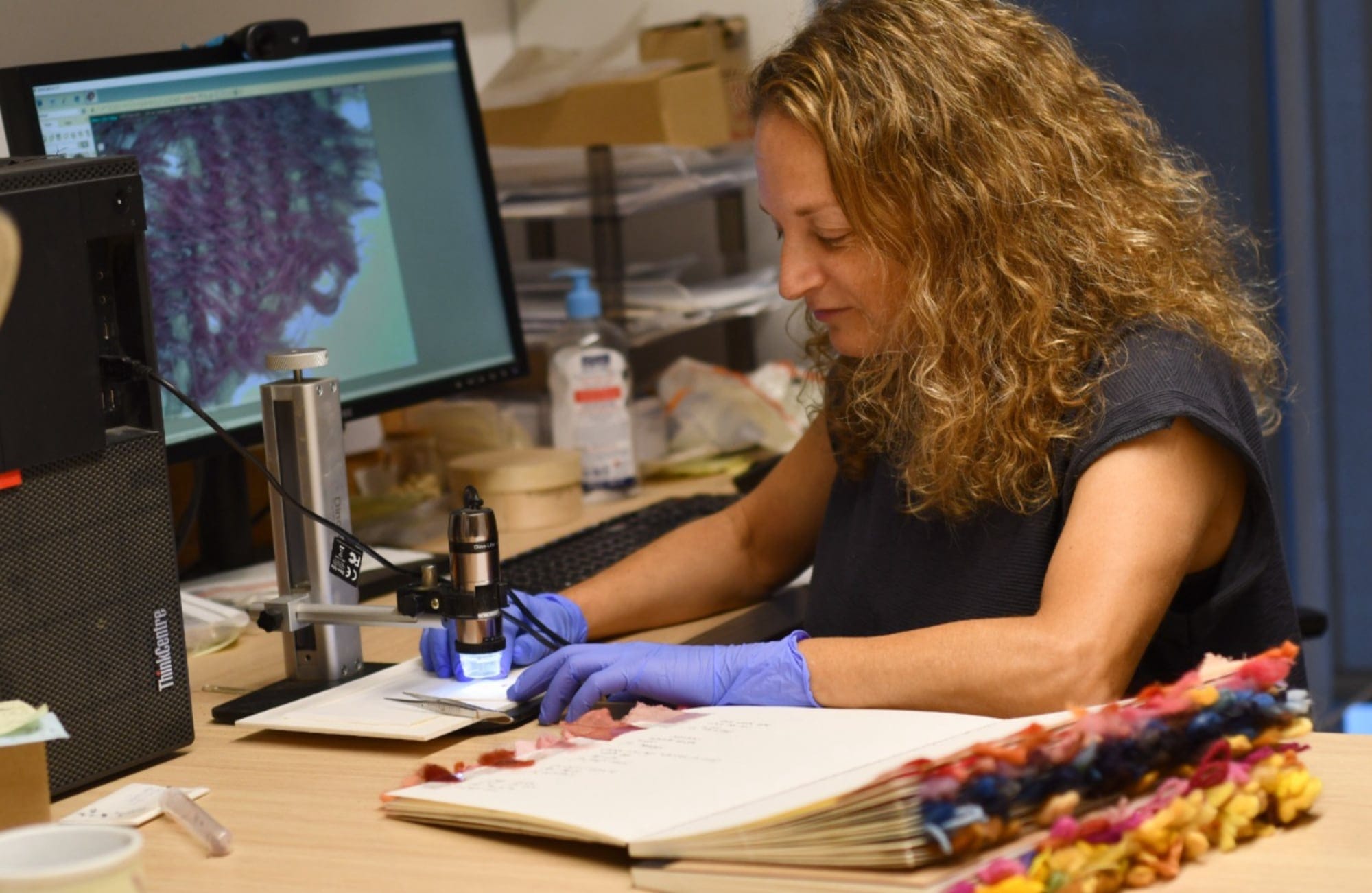 a woman with curly blond hair and purple gloves looks down through a microscope at a textile