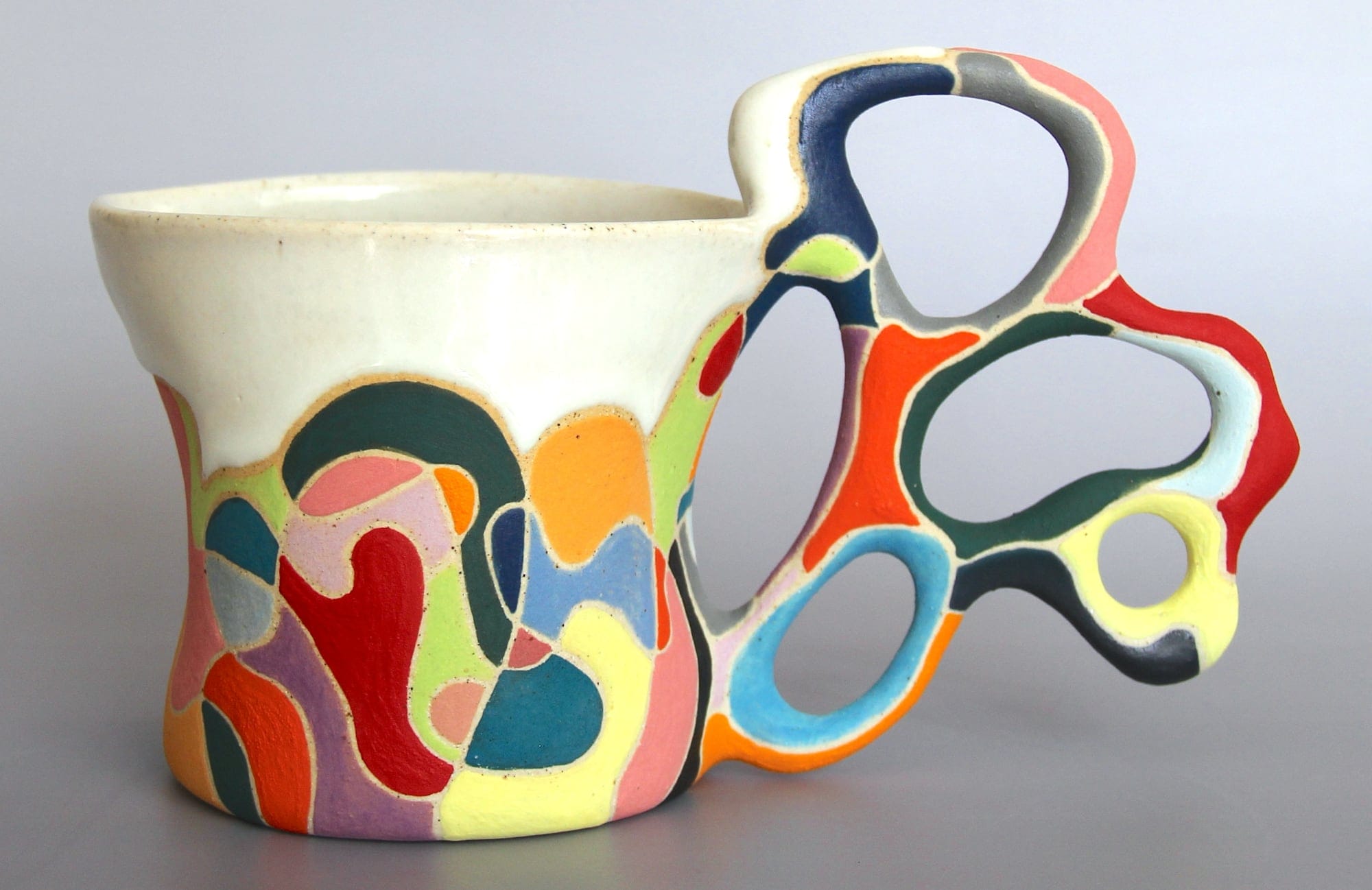 a ceramic mug with a white glaze on the top of the vessel and a vibrant patchwork below. a sprawling, biomorphic handle covered in bright covers appears to grow from the side