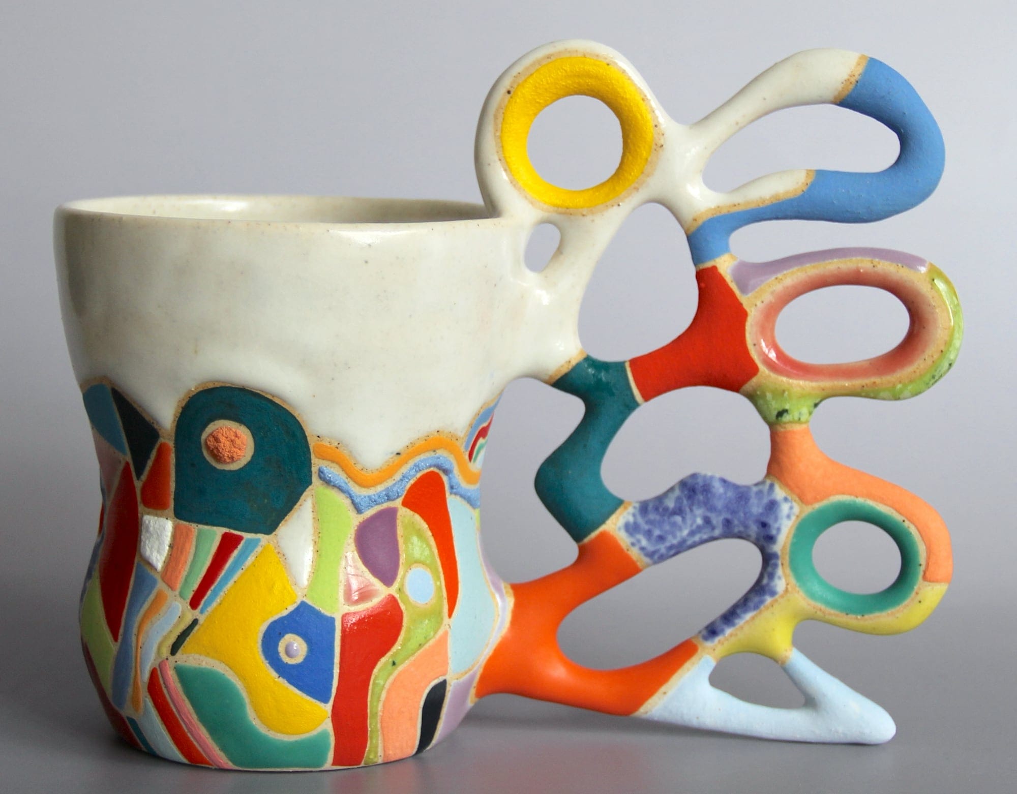 a ceramic mug with a white glaze on the top of the vessel and a vibrant patchwork below. a sprawling, biomorphic handle covered in bright covers appears to grow from the side