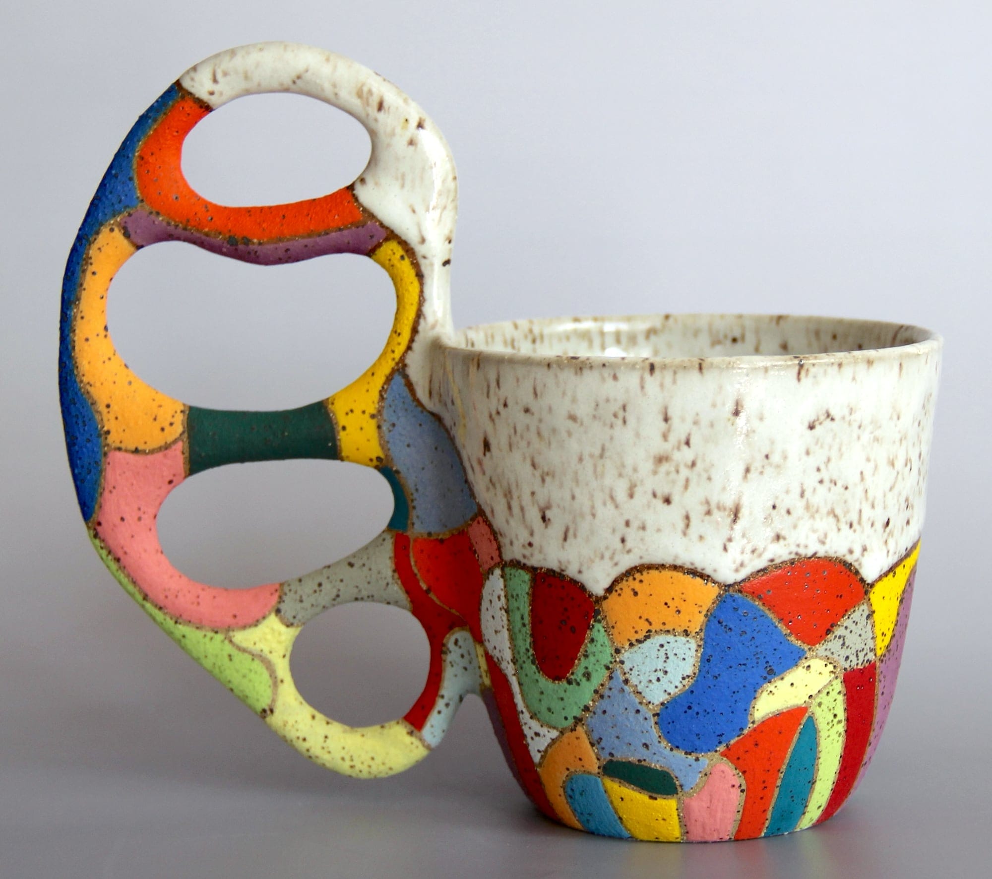 a ceramic mug with a white glaze on the top of the vessel and a vibrant patchwork below. a sprawling, biomorphic handle covered in bright covers appears to grow from the side