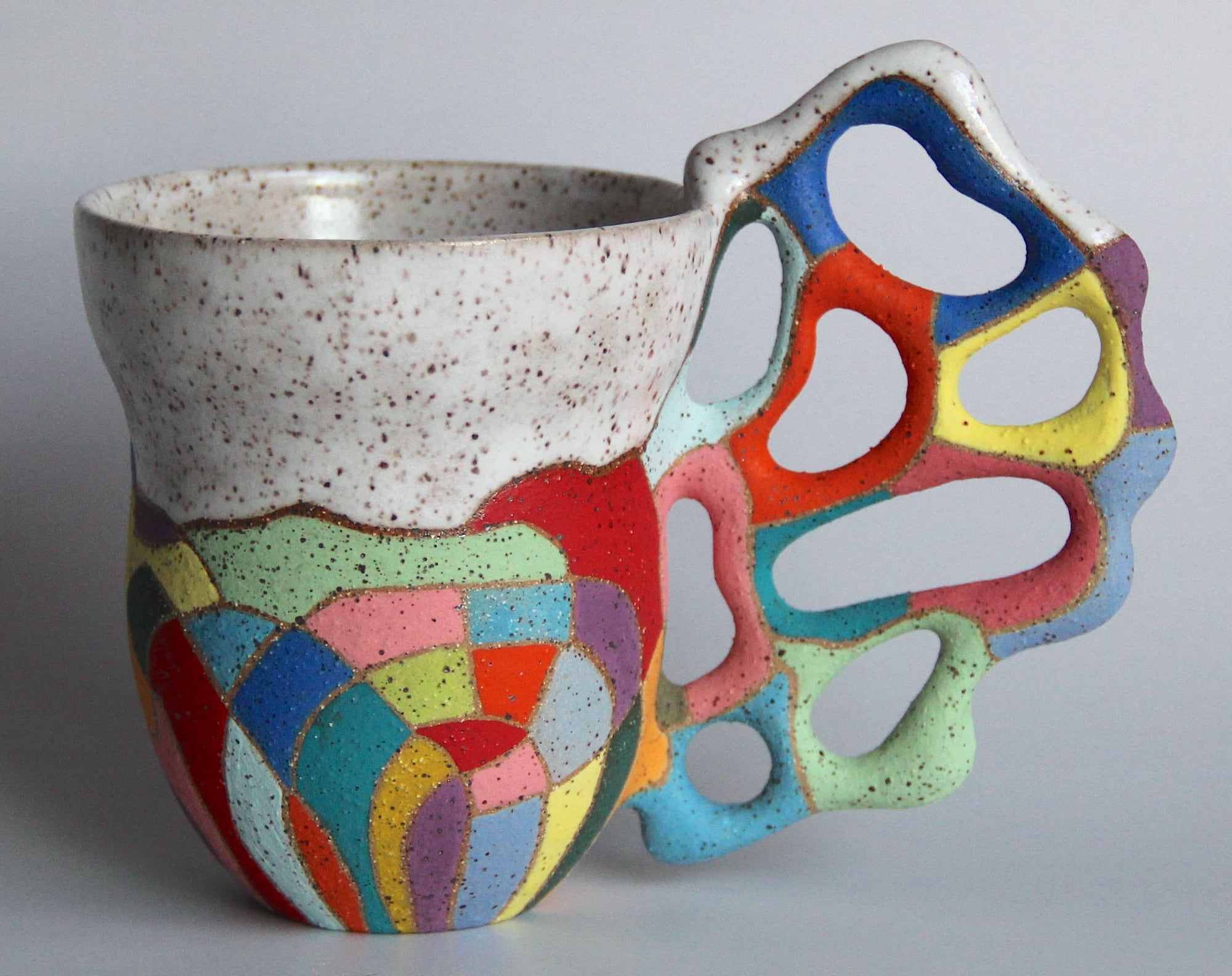 a ceramic mug with a white glaze on the top of the vessel and a vibrant patchwork below. a sprawling, biomorphic handle covered in bright covers appears to grow from the side