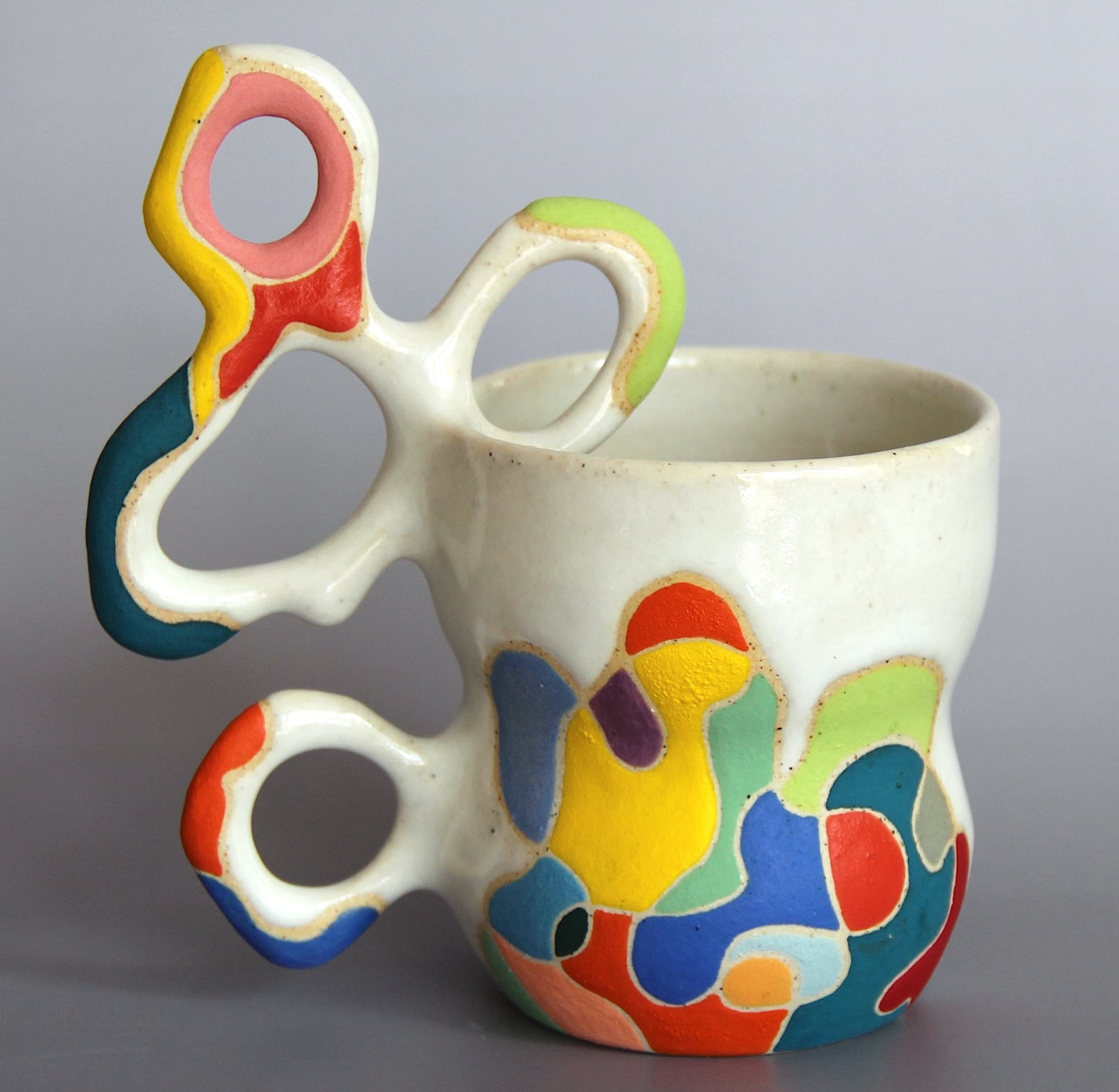 a ceramic mug with a white glaze on the top of the vessel and a vibrant patchwork below. a sprawling, biomorphic handle covered in bright covers appears to grow from the side