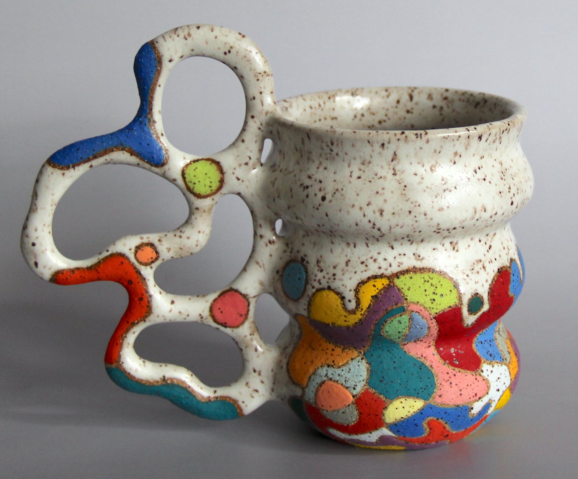 a ceramic mug with a white glaze on the top of the vessel and a vibrant patchwork below. a sprawling, biomorphic handle covered in bright covers appears to grow from the side