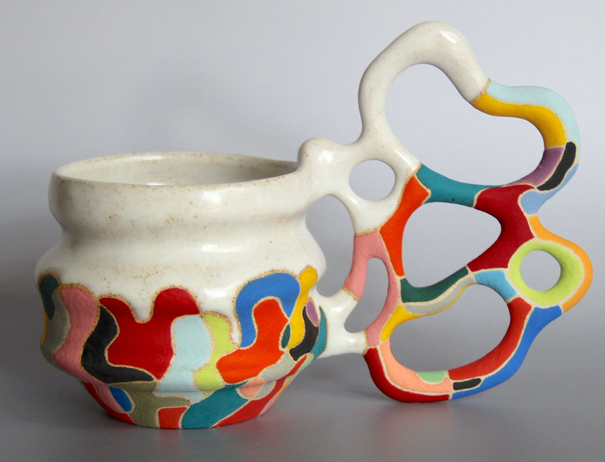 a ceramic mug with a white glaze on the top of the vessel and a vibrant patchwork below. a sprawling, biomorphic handle covered in bright covers appears to grow from the side