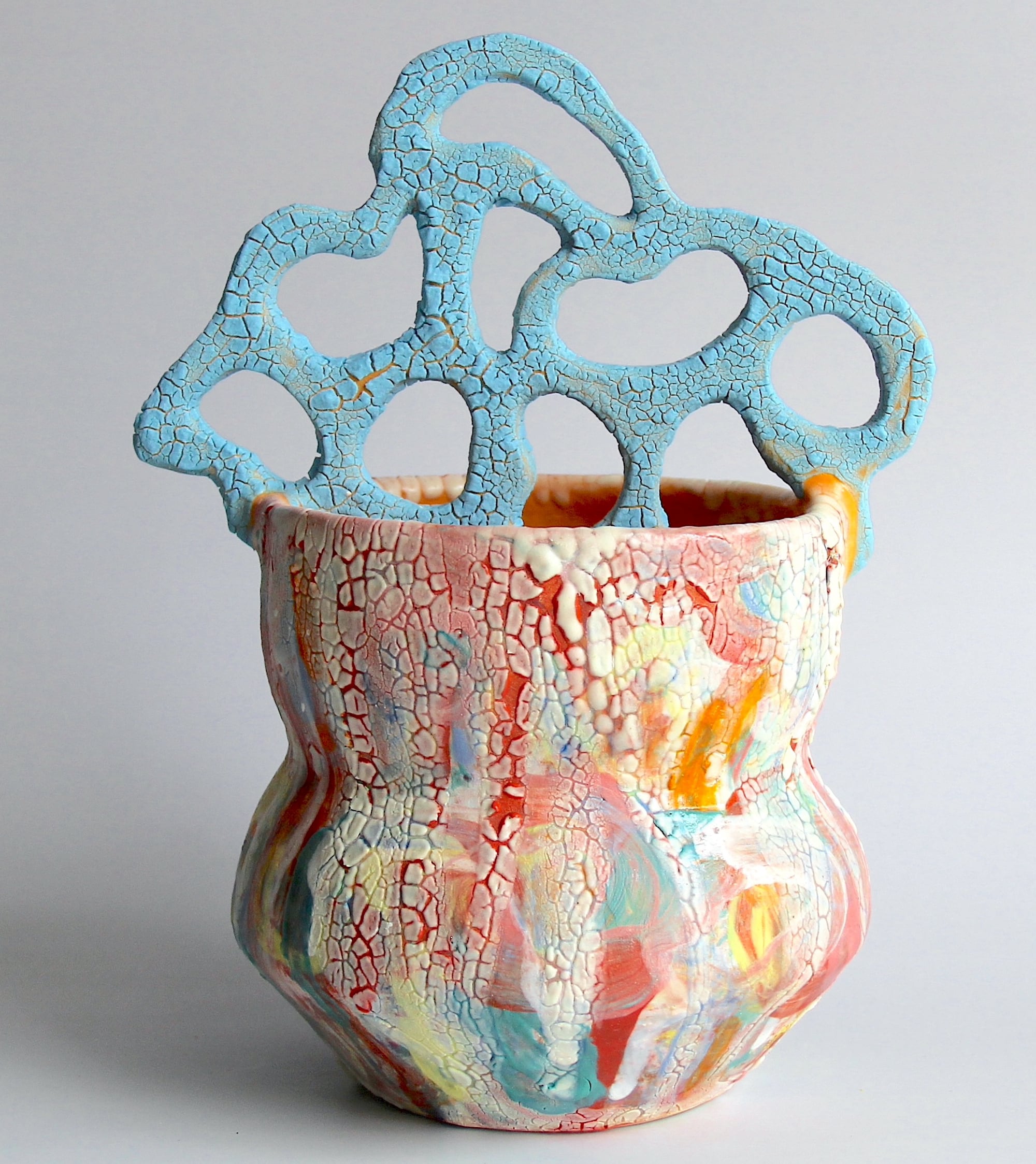 a ceramic mug with a white glaze on the top of the vessel and a vibrant patchwork below. a sprawling, biomorphic handle covered in bright covers appears to grow from inside. all is covered in a crackled glaze