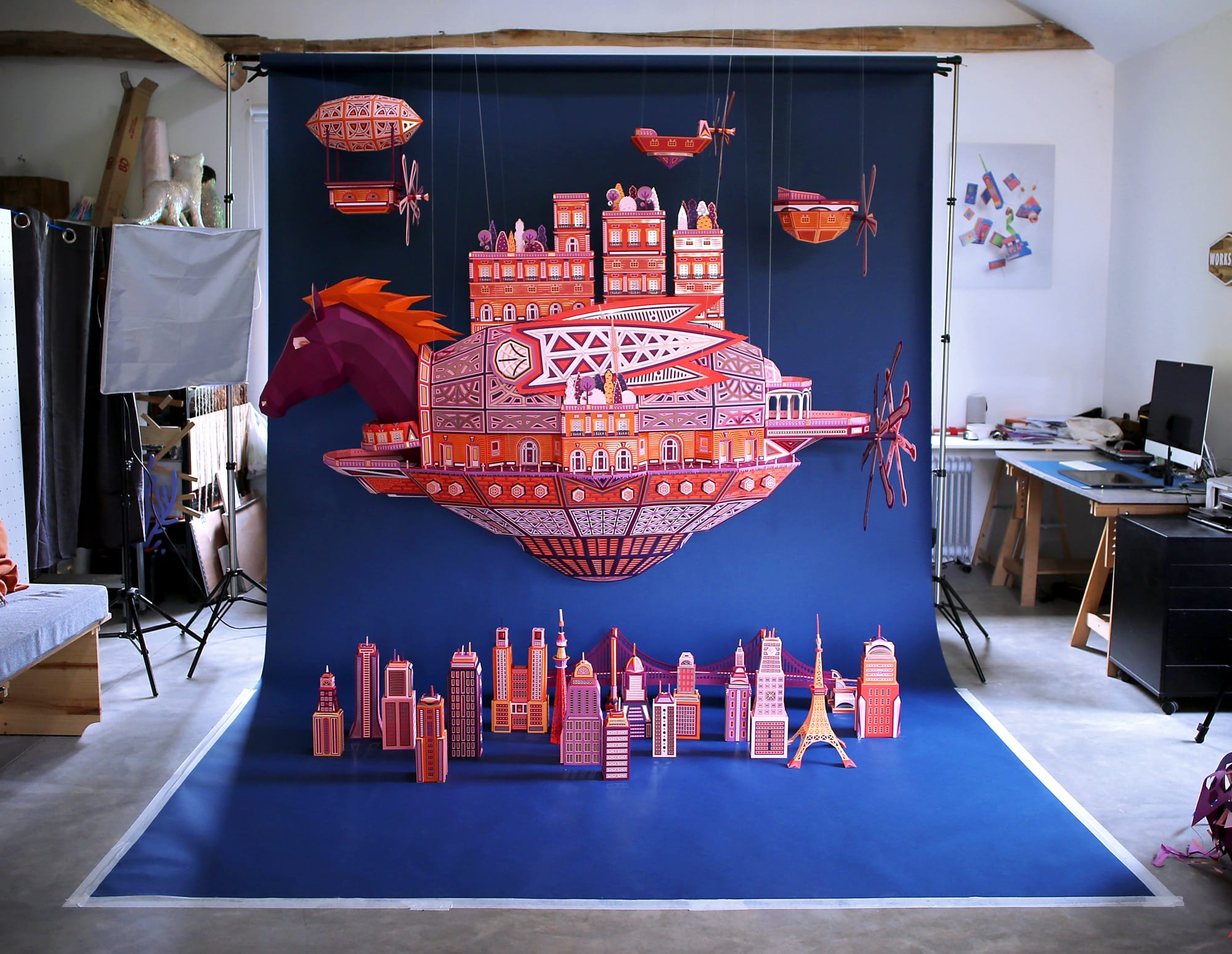 a behind the scenes shot of a massive horse head in purple and orange paper leading a flying ship with several small ships suspended from the ceiling above. a small city populates the area below on a bright blue backdrop in the artist's studio