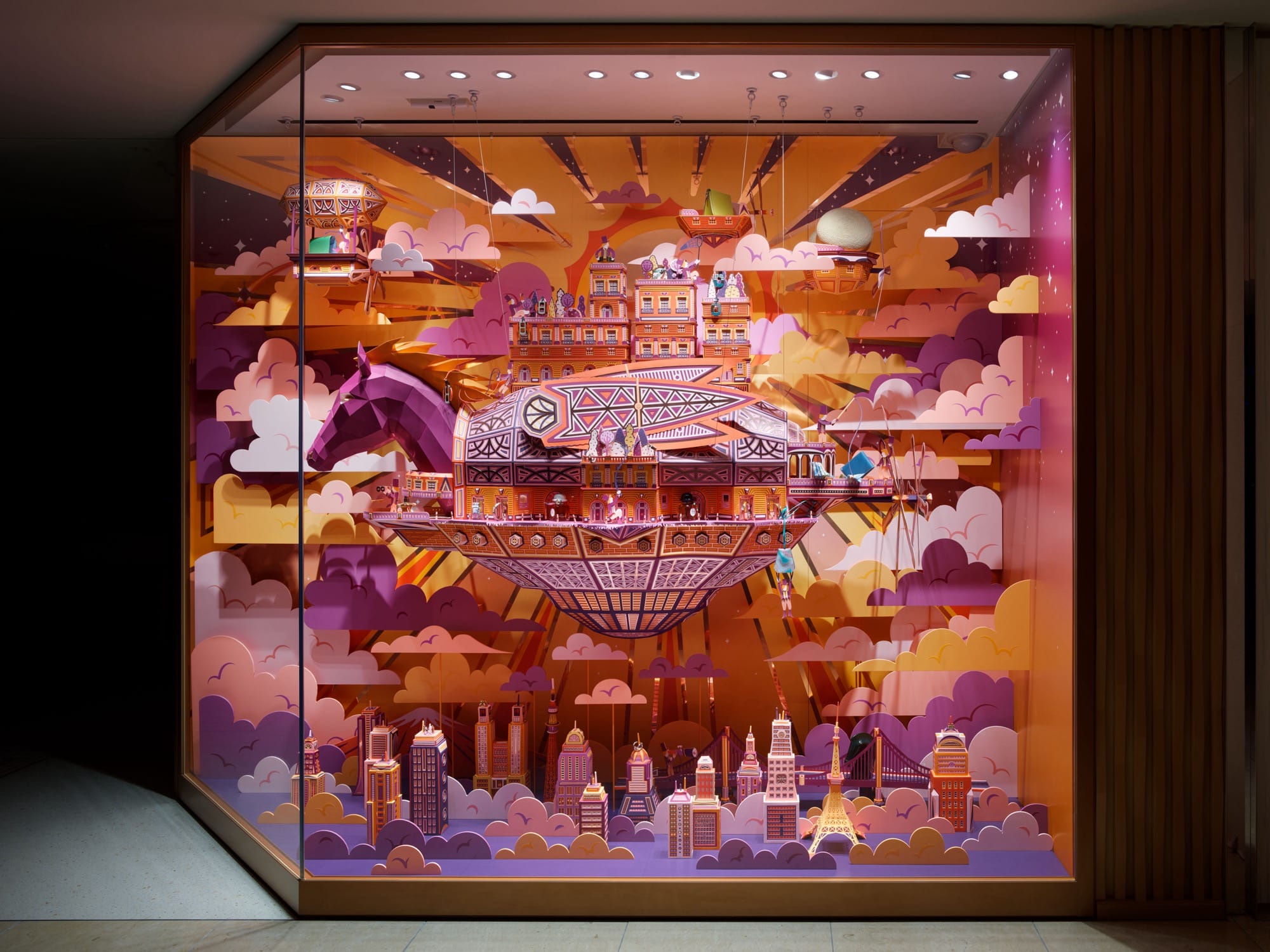 a paper installation window display featuring a purple and orange horse head leading a flying ship surrounded by clouds, suspended smaller ships, and a cityscape below