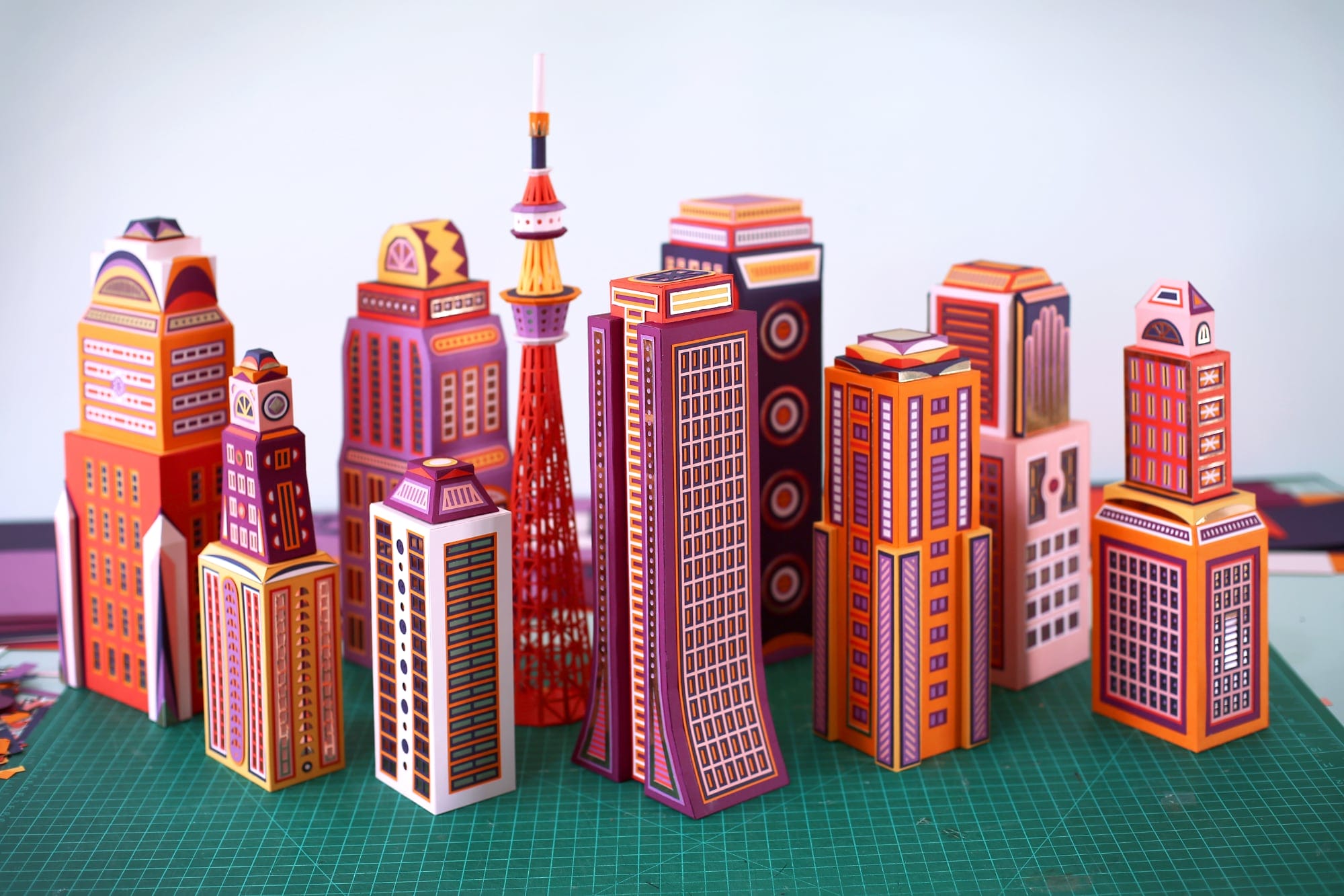 a work in progress shot on a green cutting mat of several tall paper buildings in orange, purple, pink, and red