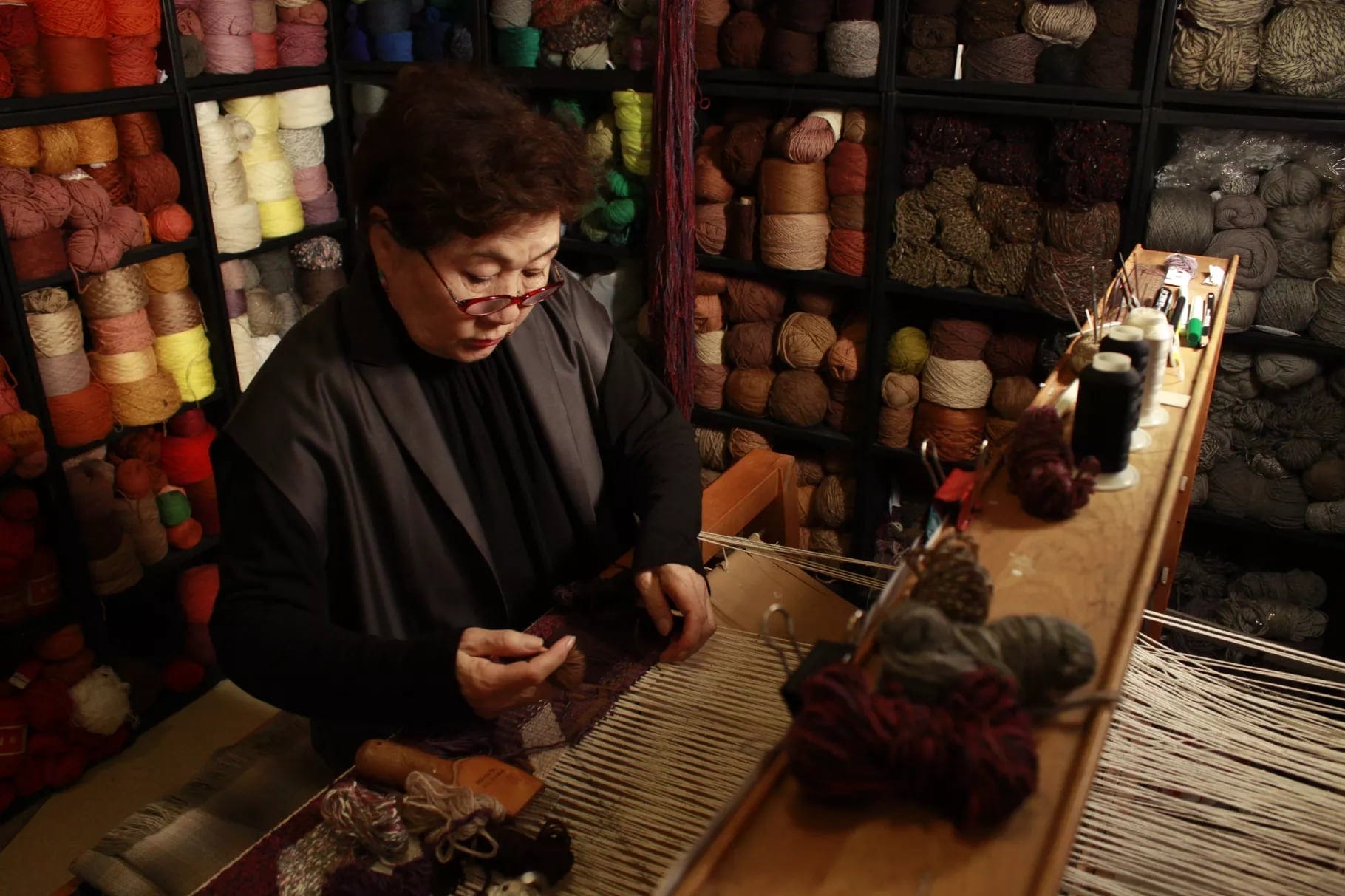 an older asian woman wearing glasses and a long sleeve black shirt pulls threads on a loom with shelves of colorful fibers behind her