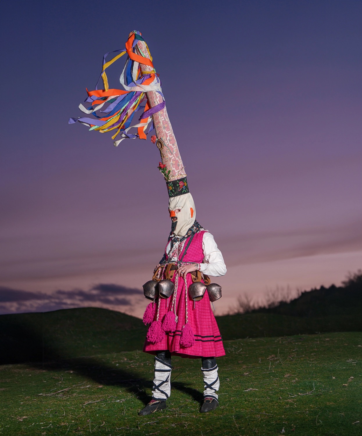 a single figure photographed at dusk with an exceedingly tall hat on, large bells around their waist, a white face covering, and a bright pink dress