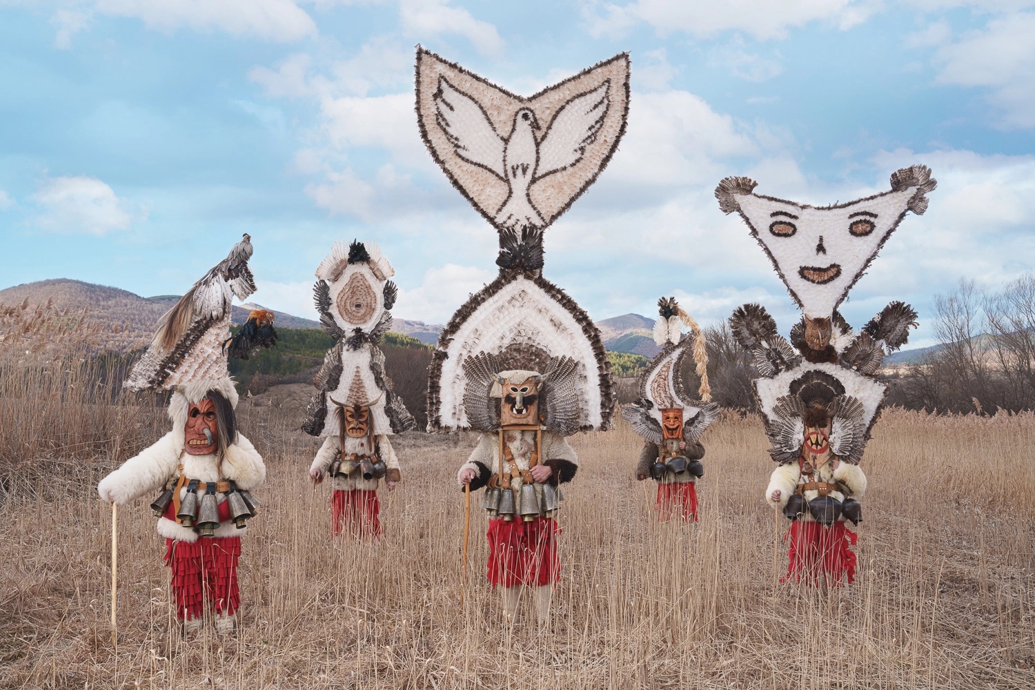 a group of five costumed figures in a field with hills in the background, wearing elaborately made, sculptural outfits with emblems of birds, faces, and other shapes