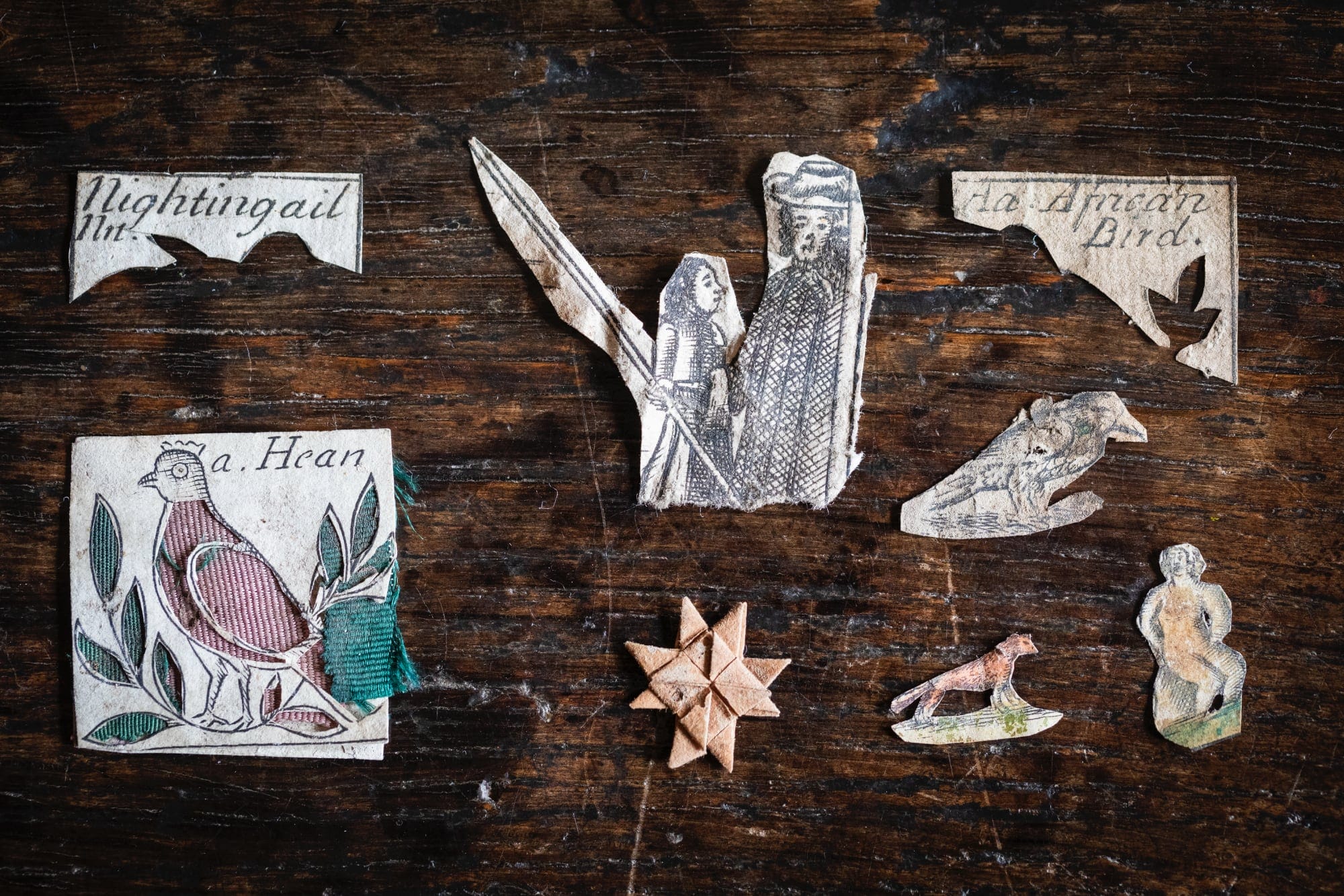 a collection of papercuts including a hen embroidered with pink and green, a couple in 17th century dress, a folded star, a fox, a bird, a woman on grass, and two corner texts reading "nightingail" and "an african bird" on a wooden surface