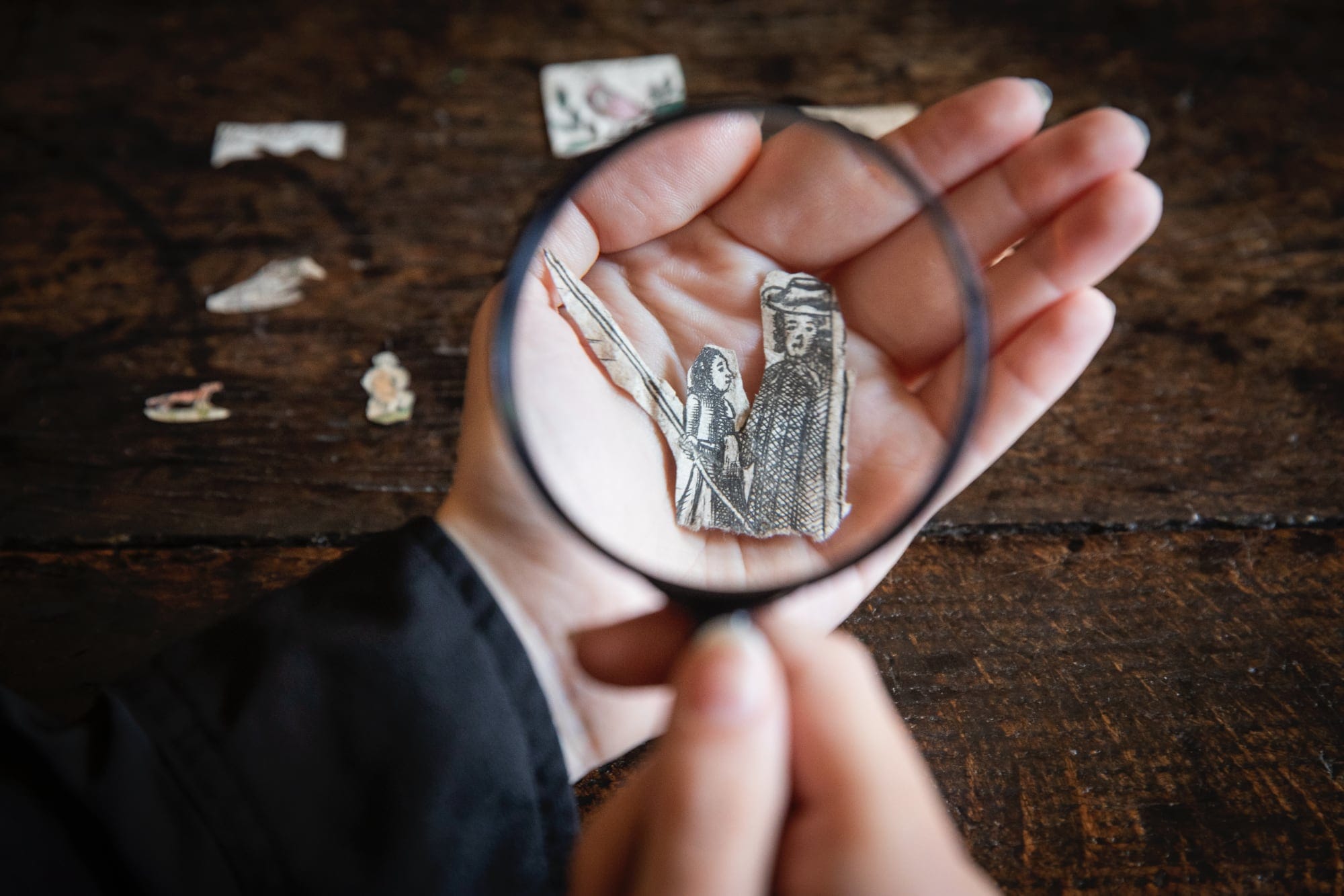 a white hand holding a papercut of a couple in 17th century dress that's magnified through a glass