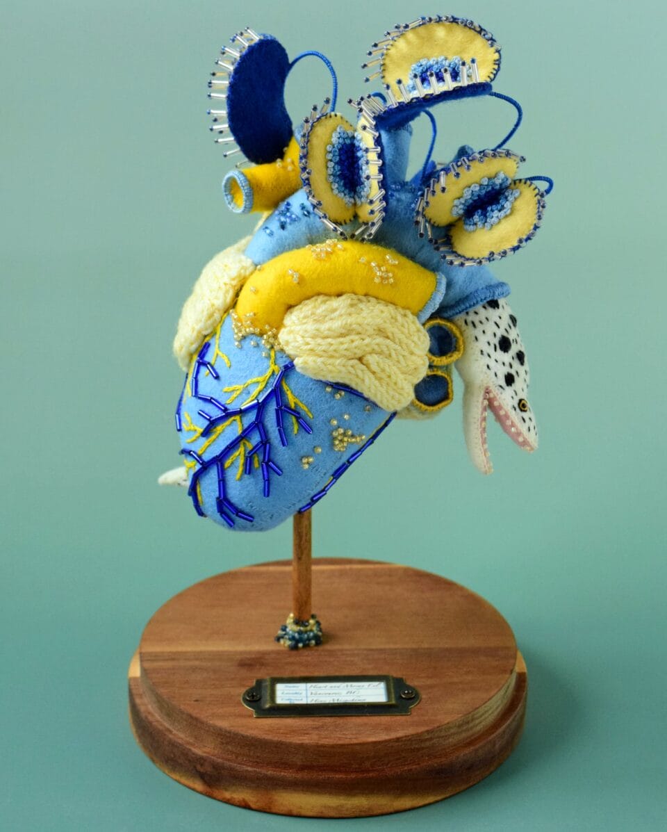 a black spotted white eel emerges with an open mouth from a blue aorta on a beaded blue heart. the sculpture features venus fly traps in blue and yellow at the top and stands on a wooden pedestal